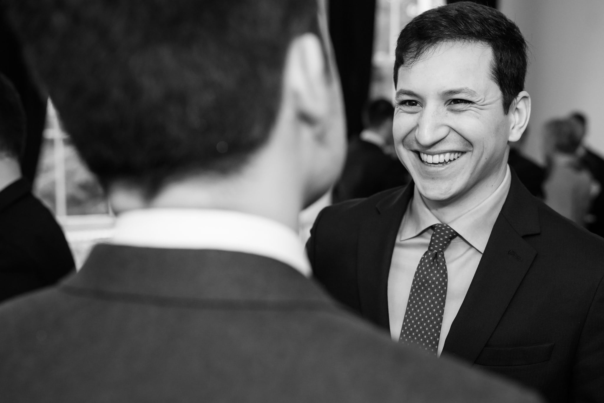 A man in a suit and tie smiles while speaking to another person, whose back is to the camera, at a formal event in Surrey.
