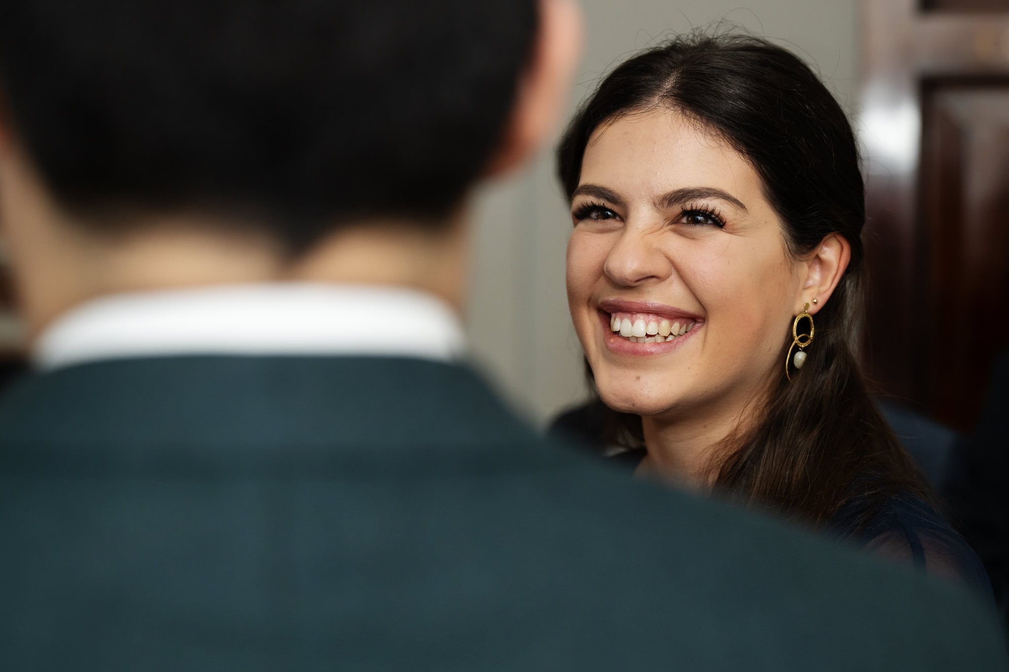 A woman with long brown hair and wearing earrings smiles while looking at someone in the foreground, capturing a joyful moment at an Addington Palace wedding.