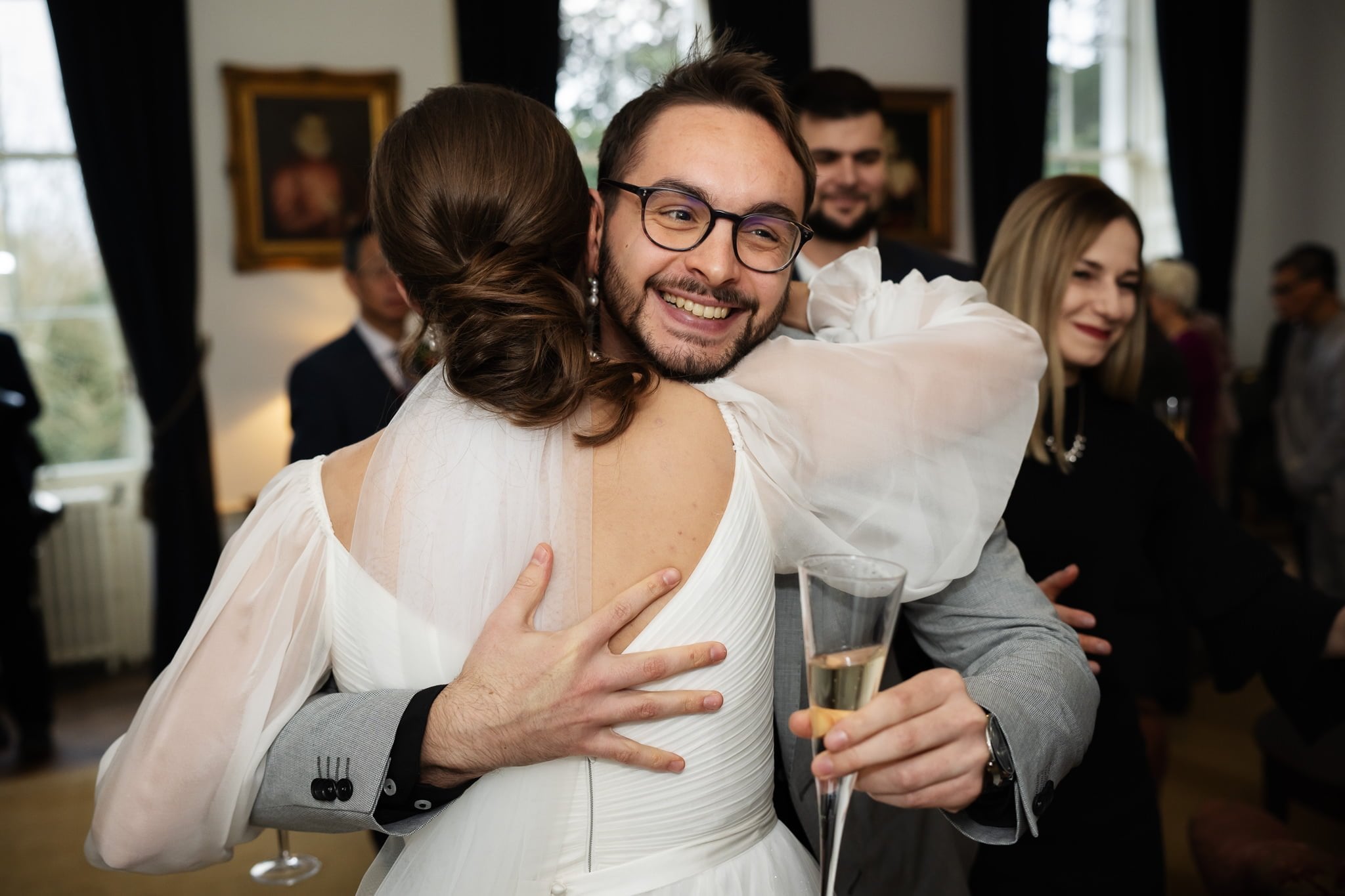A groom wearing glasses and holding a champagne flute hugs the bride while smiling at a wedding gathering indoors. Other guests are visible in the background, capturing the joy of their Addington Palace wedding in Surrey through skilled Addington Palace wedding photography.