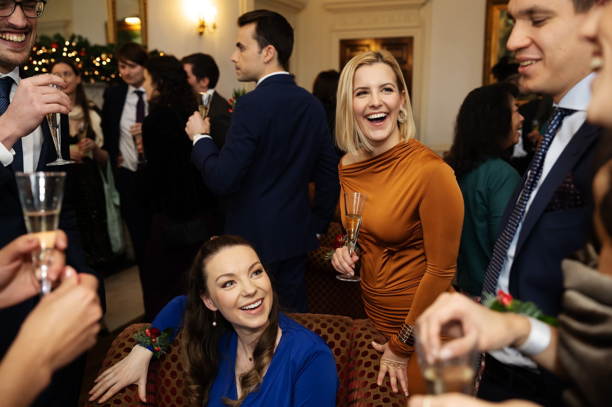 A group of people, dressed in formal attire, are socializing and holding champagne flutes at an indoor wedding gathering. Some are smiling and engaged in conversation, enjoying the elegant ambiance at the Addington Palace wedding in Surrey.
