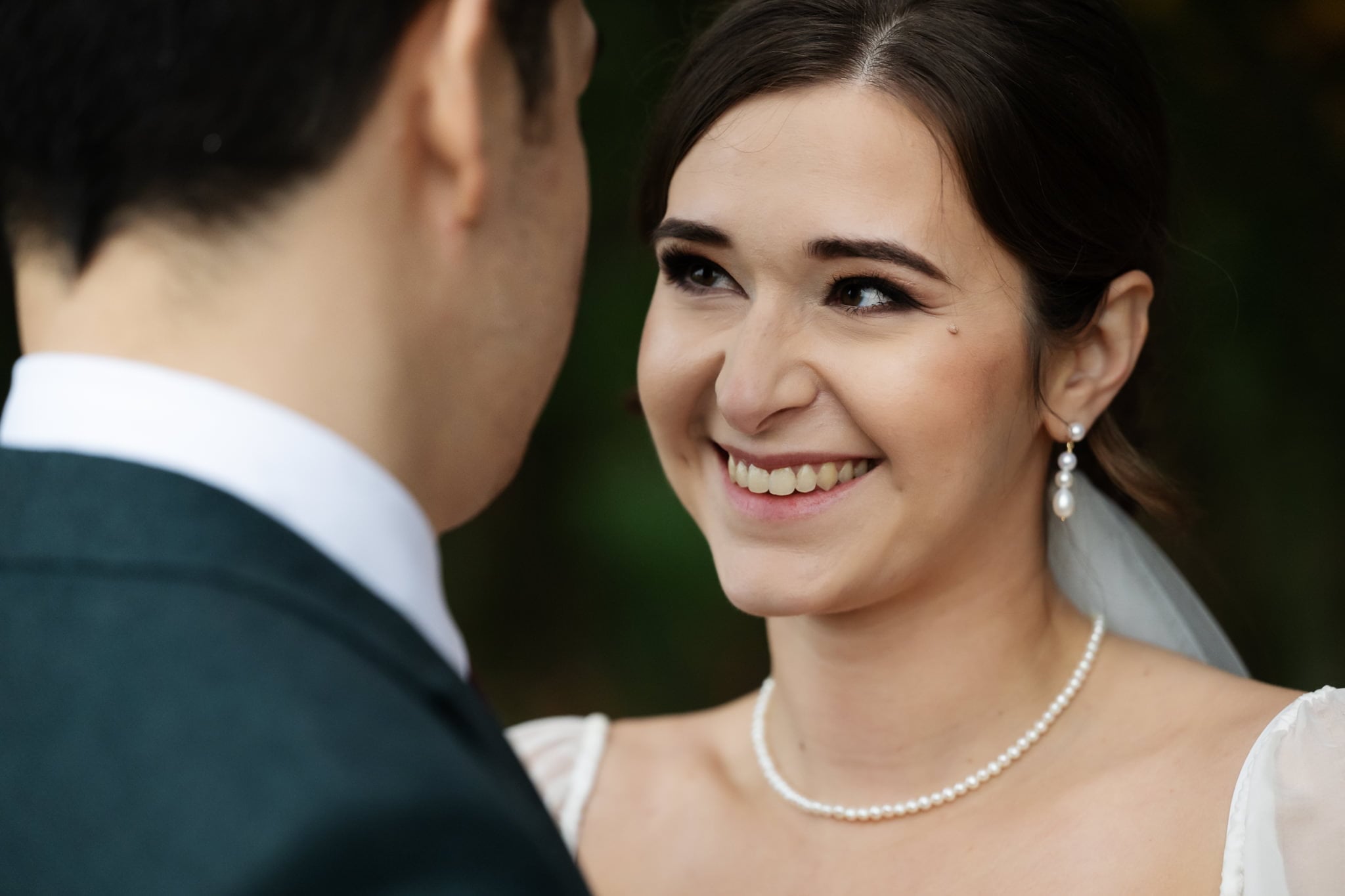A bride smiling at a groom while they face each other, with the bride wearing a pearl necklace and earrings, captured beautifully by an Addington Palace wedding photographer.
