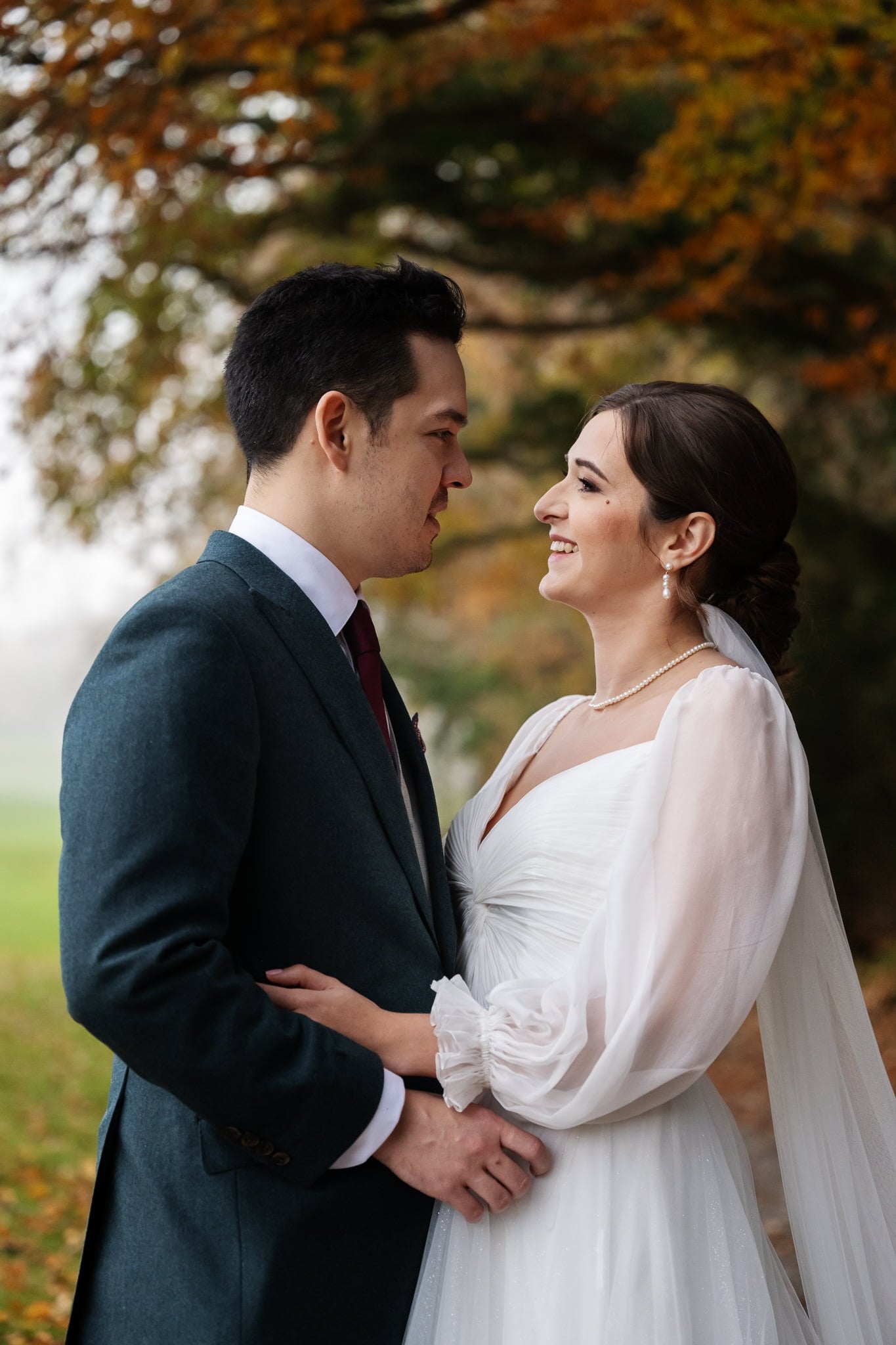 A bride and groom stand outdoors in wedding attire, looking at each other and smiling, with autumn leaves in the background—captured perfectly by their Addington Palace wedding photographer.