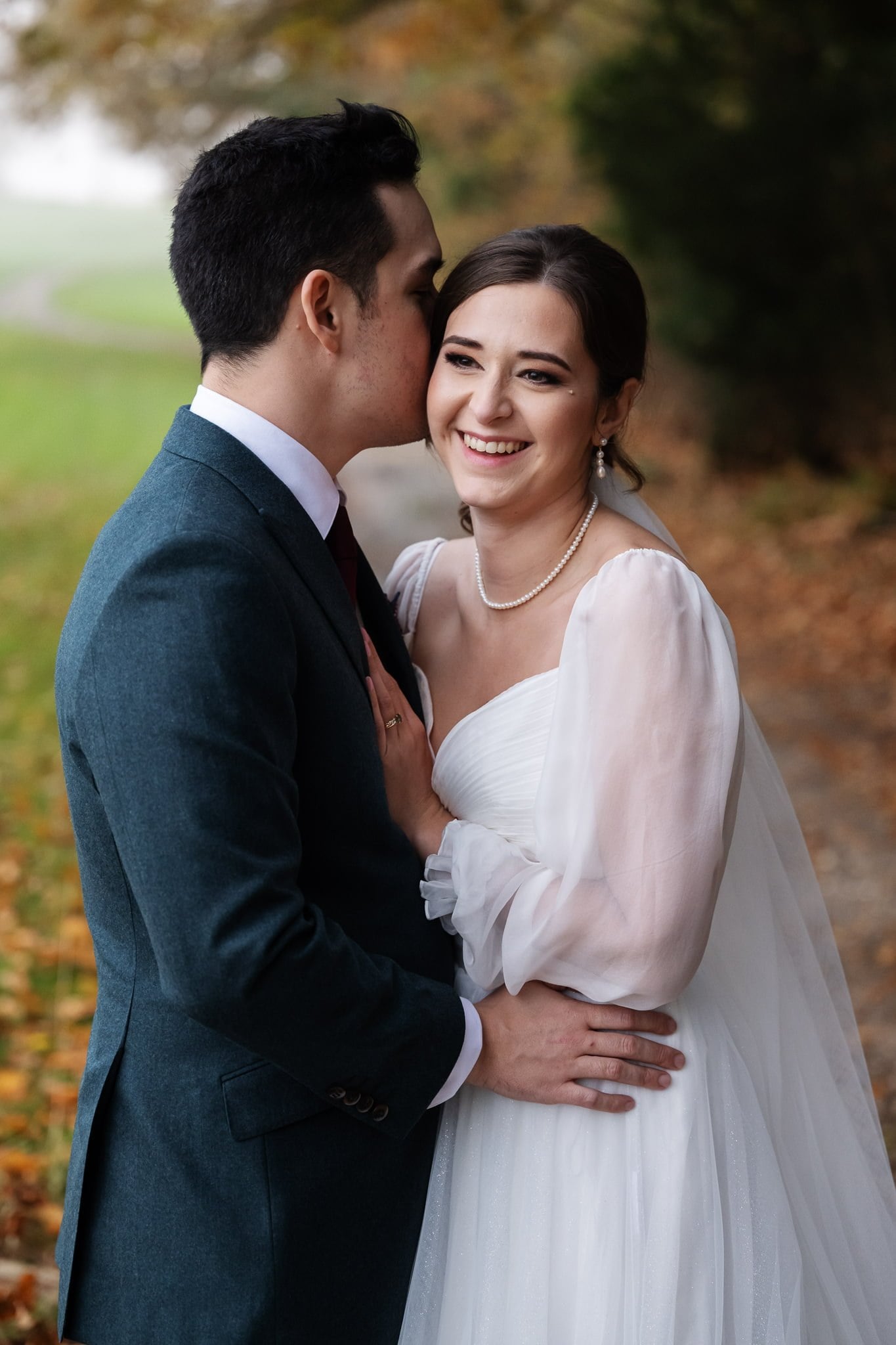 A groom in a green suit kisses a smiling bride in a white dress. They stand close together outdoors with autumn leaves scattered on the ground, celebrating their Addington Palace wedding in beautiful Surrey.