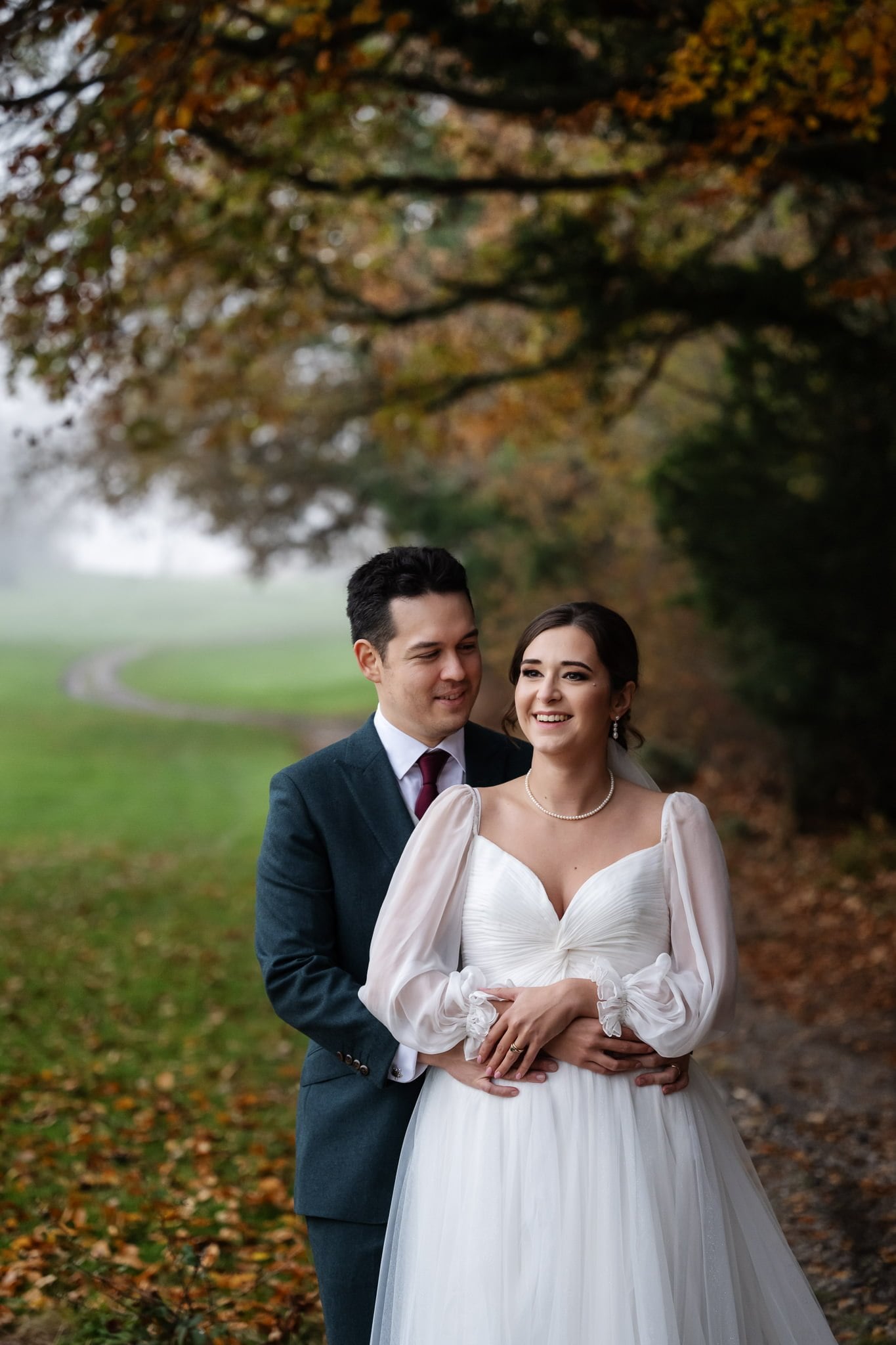 A couple stands outdoors, with the groom in a dark suit embracing the bride in a white wedding dress. The background features a misty pathway lined with autumn trees. Captured by an Addington Palace wedding photographer, this Surrey scene exudes timeless romance.