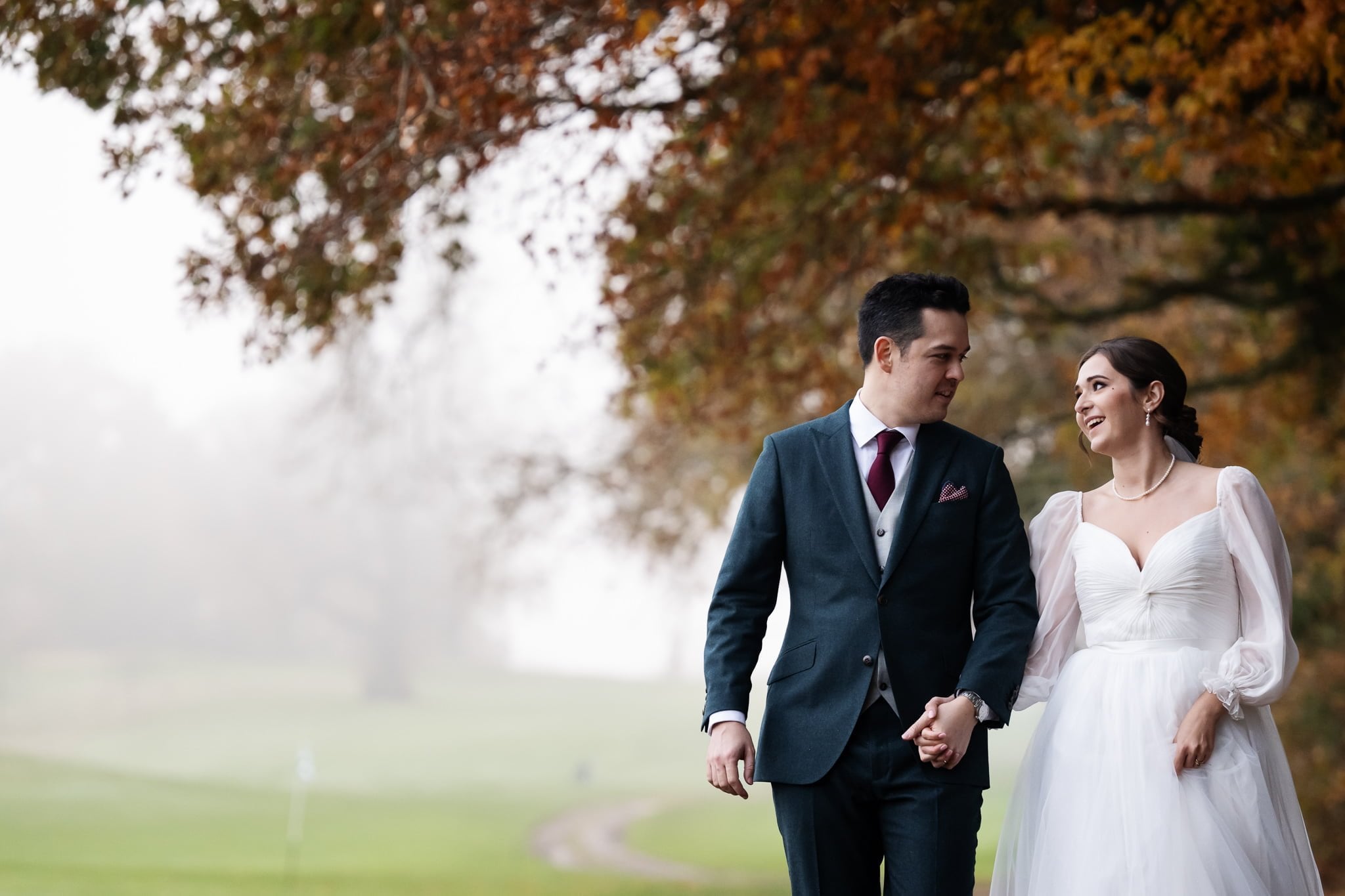 A couple dressed in wedding attire, holding hands and walking outdoors under autumn trees at Addington Palace. The groom is in a dark suit, and the bride is in a white dress. The background is misty with green grass visible, capturing the essence of their Surrey love story.