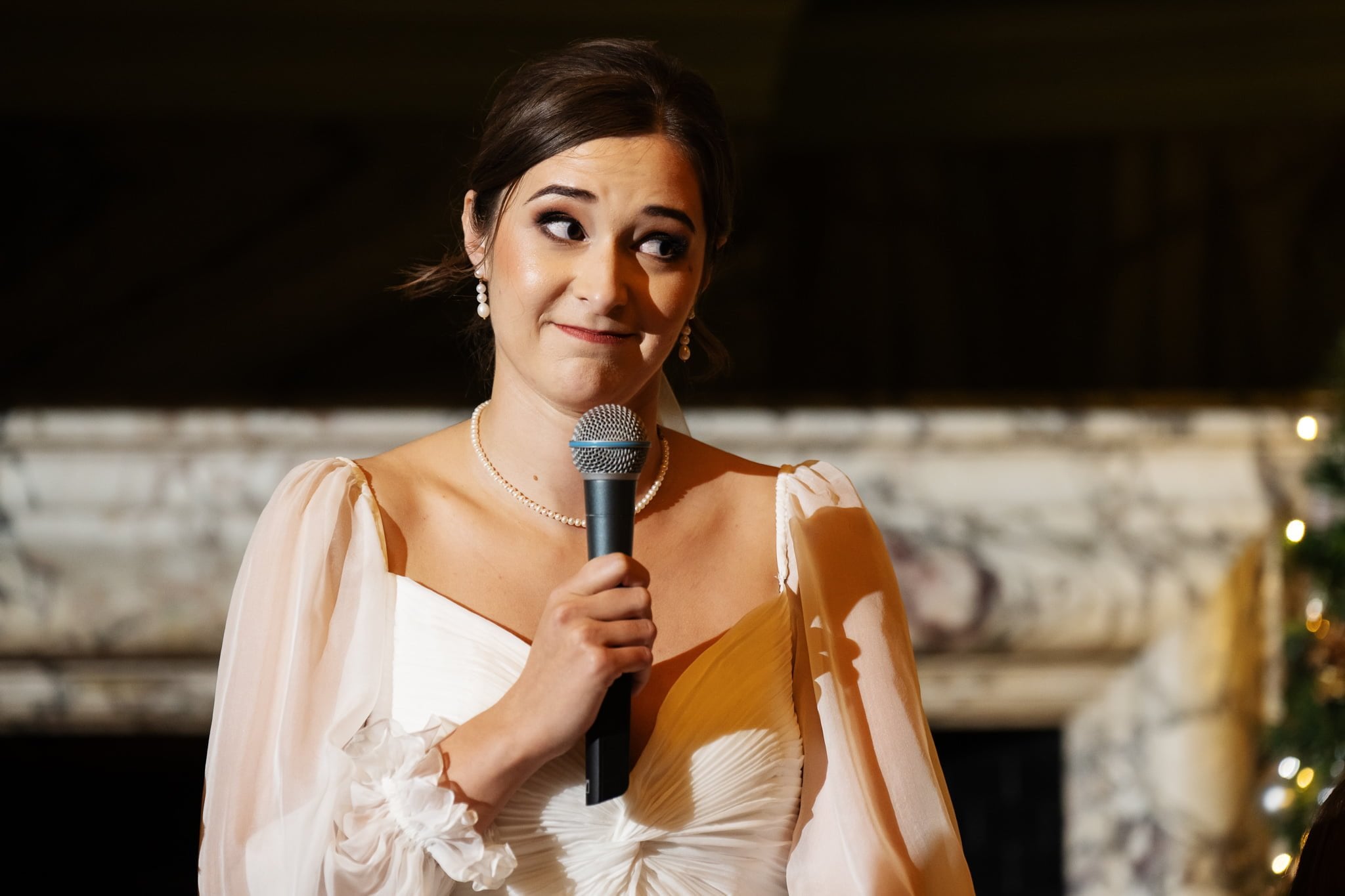 A woman in a white dress holds a microphone and looks to the side, standing in front of a marble backdrop, capturing the elegance of an Addington Palace wedding.