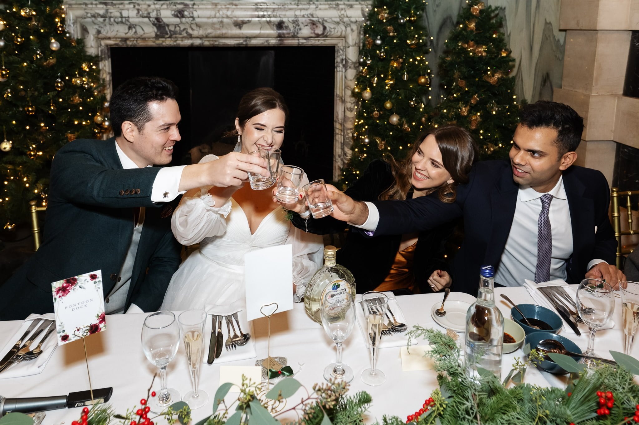 A group of four people dressed in formal attire are toasting glasses at a decorated dining table, set for a celebration with holiday-themed decorations and a festive atmosphere, reminiscent of an Addington Palace wedding photographer capturing the perfect moment.