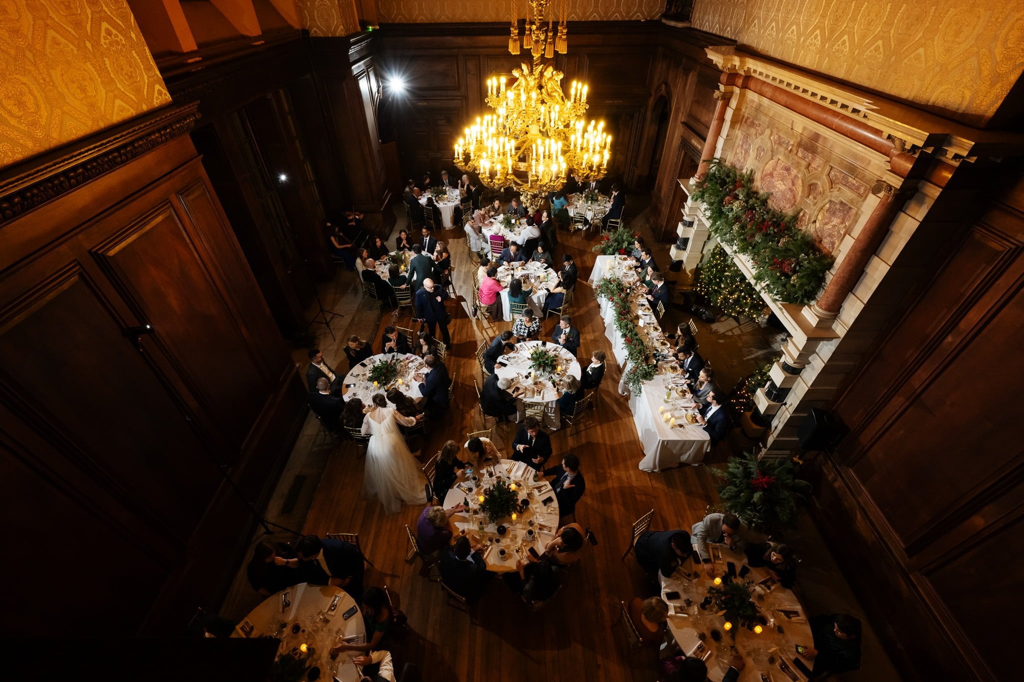 A view from above of a formal banquet hall filled with round tables, guests dining, and a large chandelier hanging in the center, featuring elegant wood paneling and decorative greenery on the fireplace mantle—perfect for capturing moments as an Addington Palace wedding photographer in Surrey.