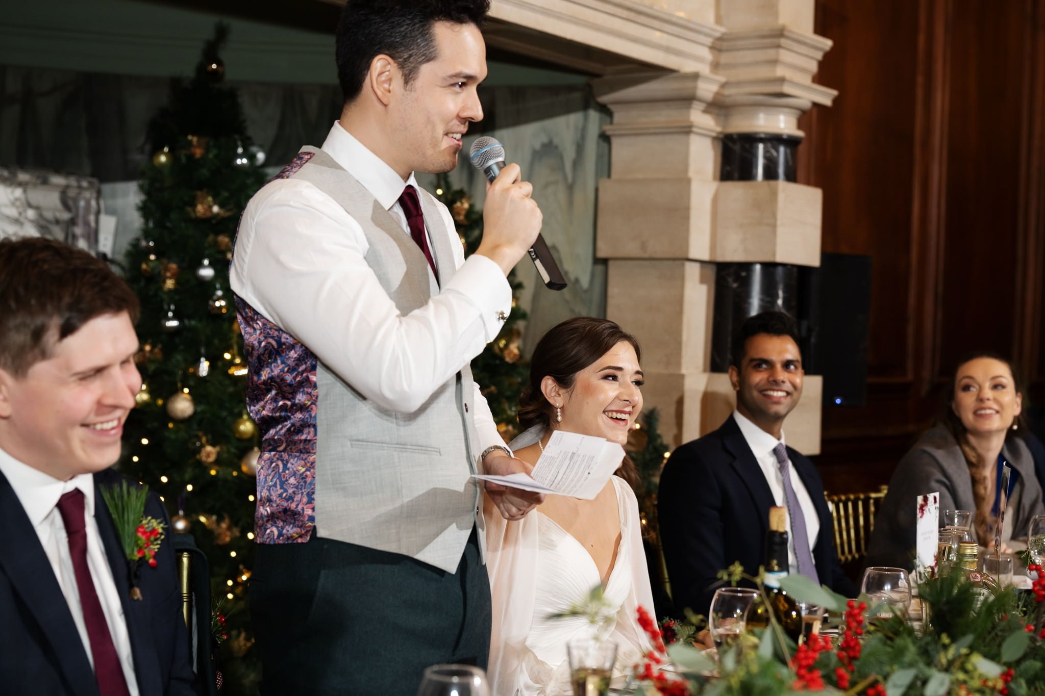 A man in a formal vest speaks into a microphone at a decorated event table, while a woman in a white dress and other attendees listen and smile. A beautifully lit Christmas tree adorns the background, capturing the enchanting atmosphere of an Addington Palace wedding.