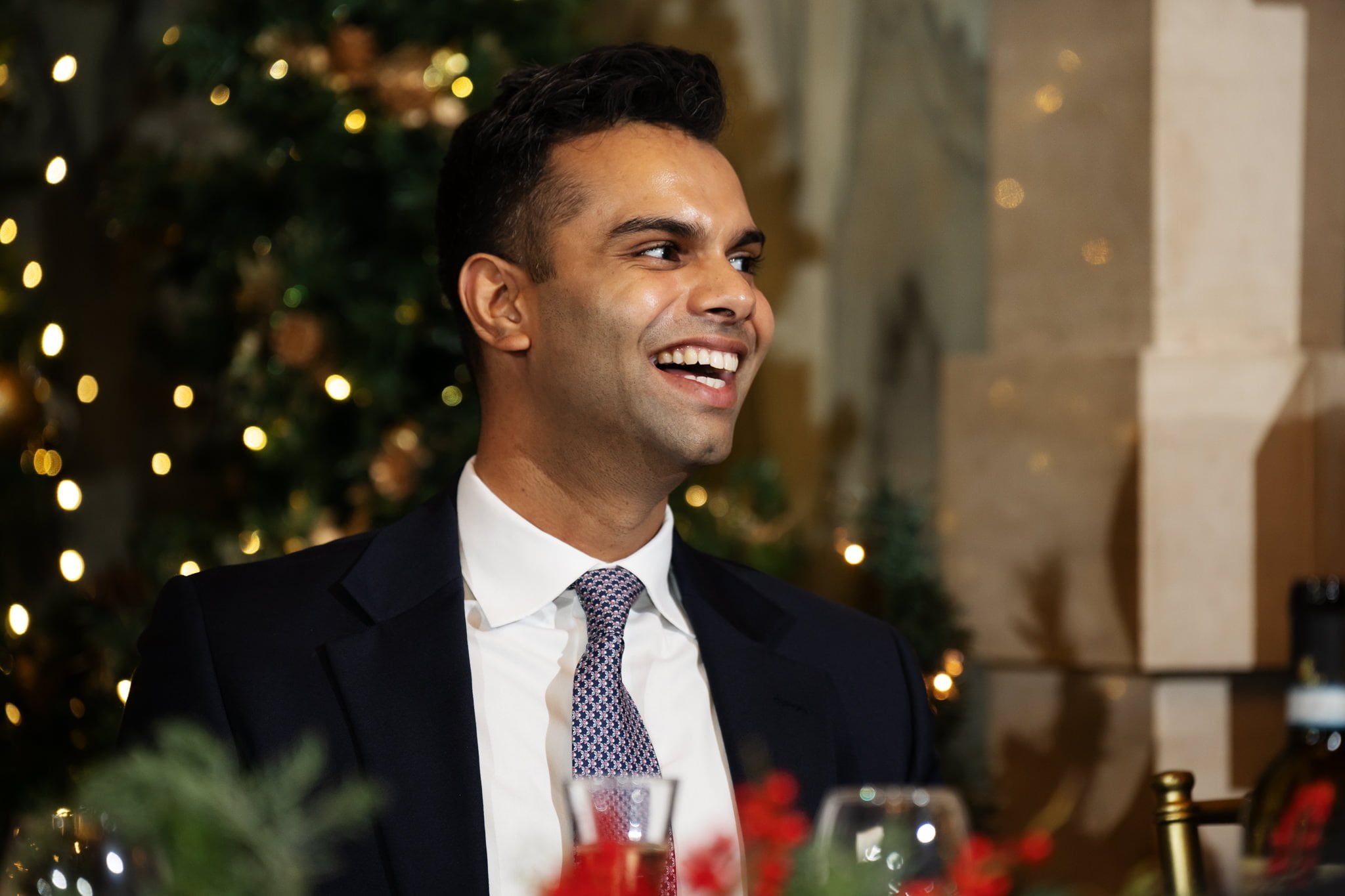 A man in a suit and tie is sitting and smiling in a festive setting with decorative lights and greenery in the background, reminiscent of a joyous celebration captured by an Addington Palace wedding photographer.