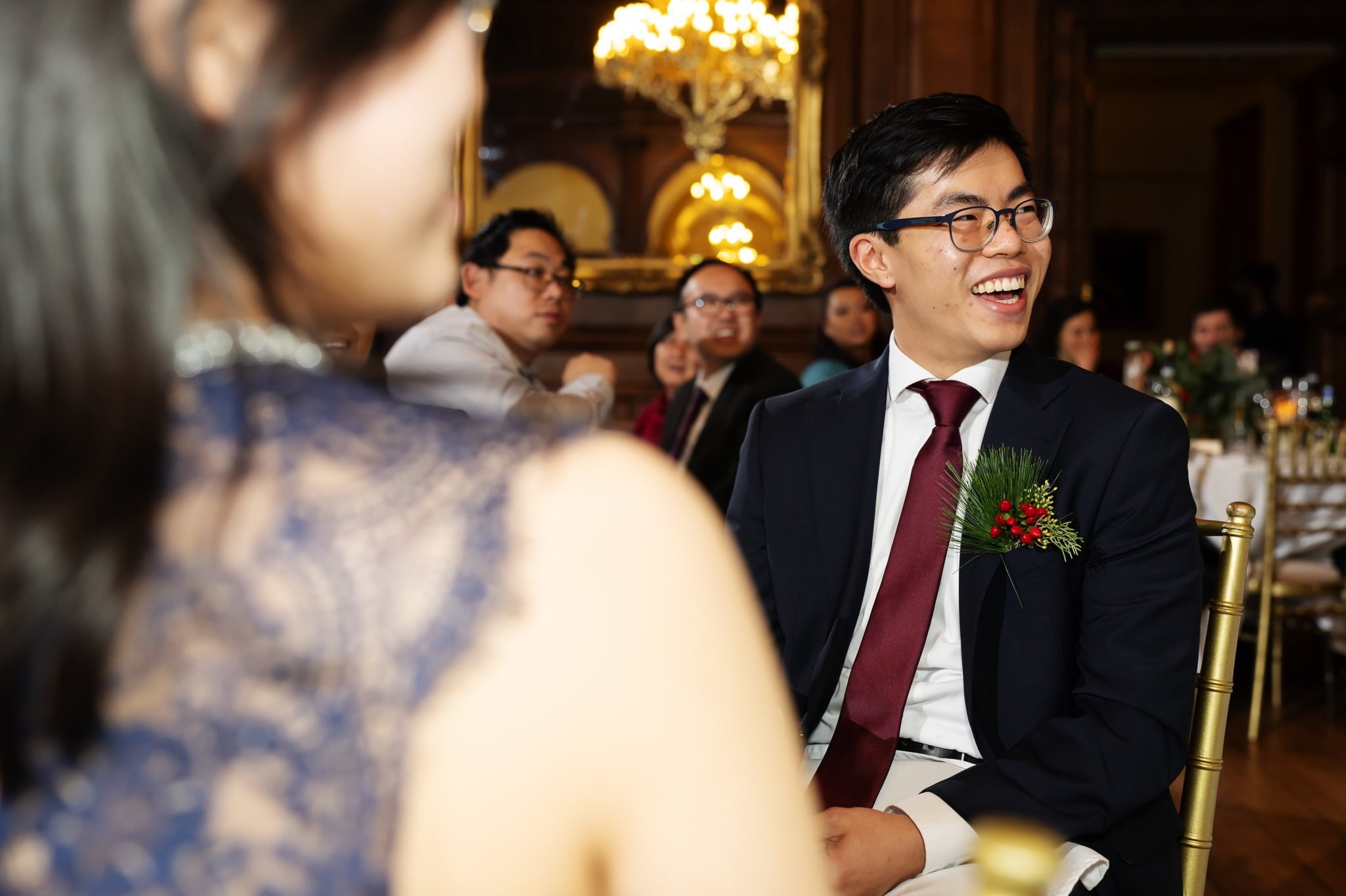 A man in a suit and tie with a red boutonniere smiles while seated at a formal wedding event, surrounded by other attendees. The moment is beautifully captured by an Addington Palace wedding photographer.
