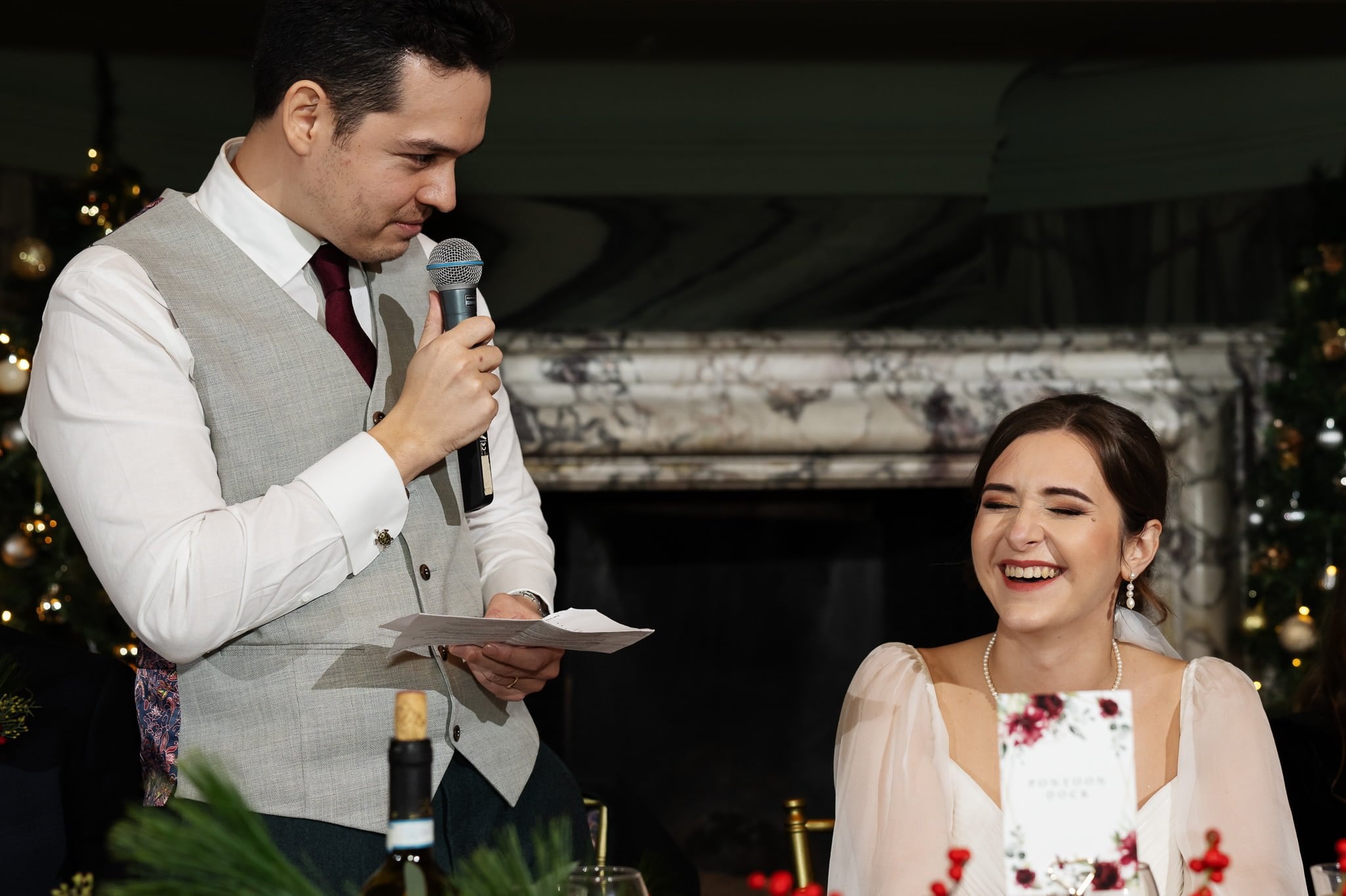 A man in a suit and tie is speaking into a microphone while holding a piece of paper at an Addington Palace wedding. A woman in a white dress is seated, smiling and looking at him. They are indoors, with a decorated background.