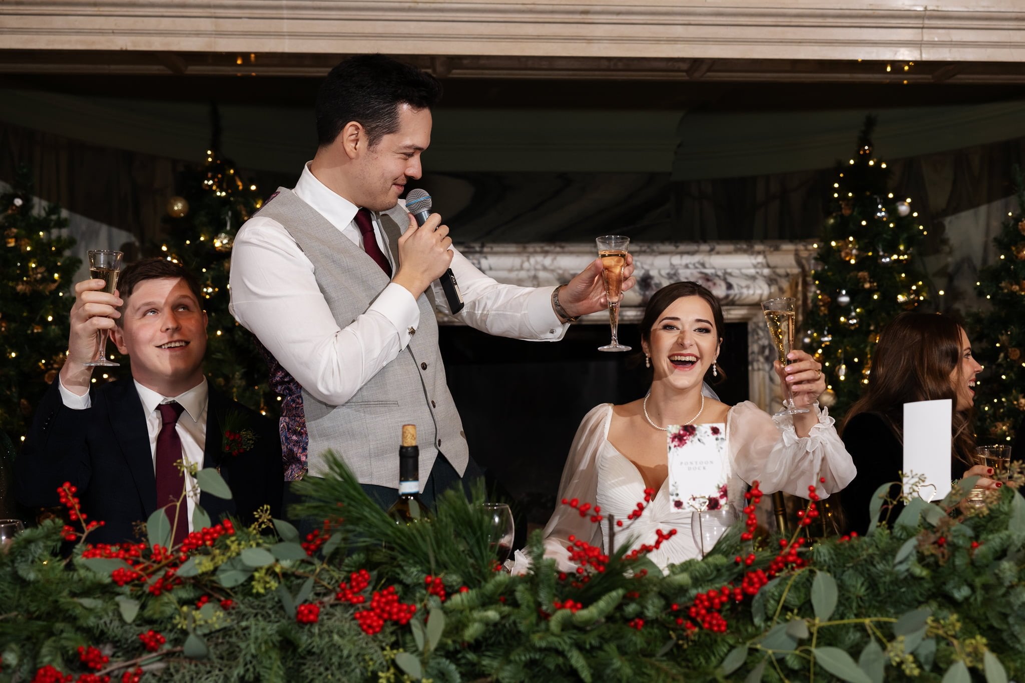 A man in a vest with a microphone and a woman in a white dress raise toasts at an Addington Palace wedding, while another man in a suit and a woman in a dark dress also join in the celebration.