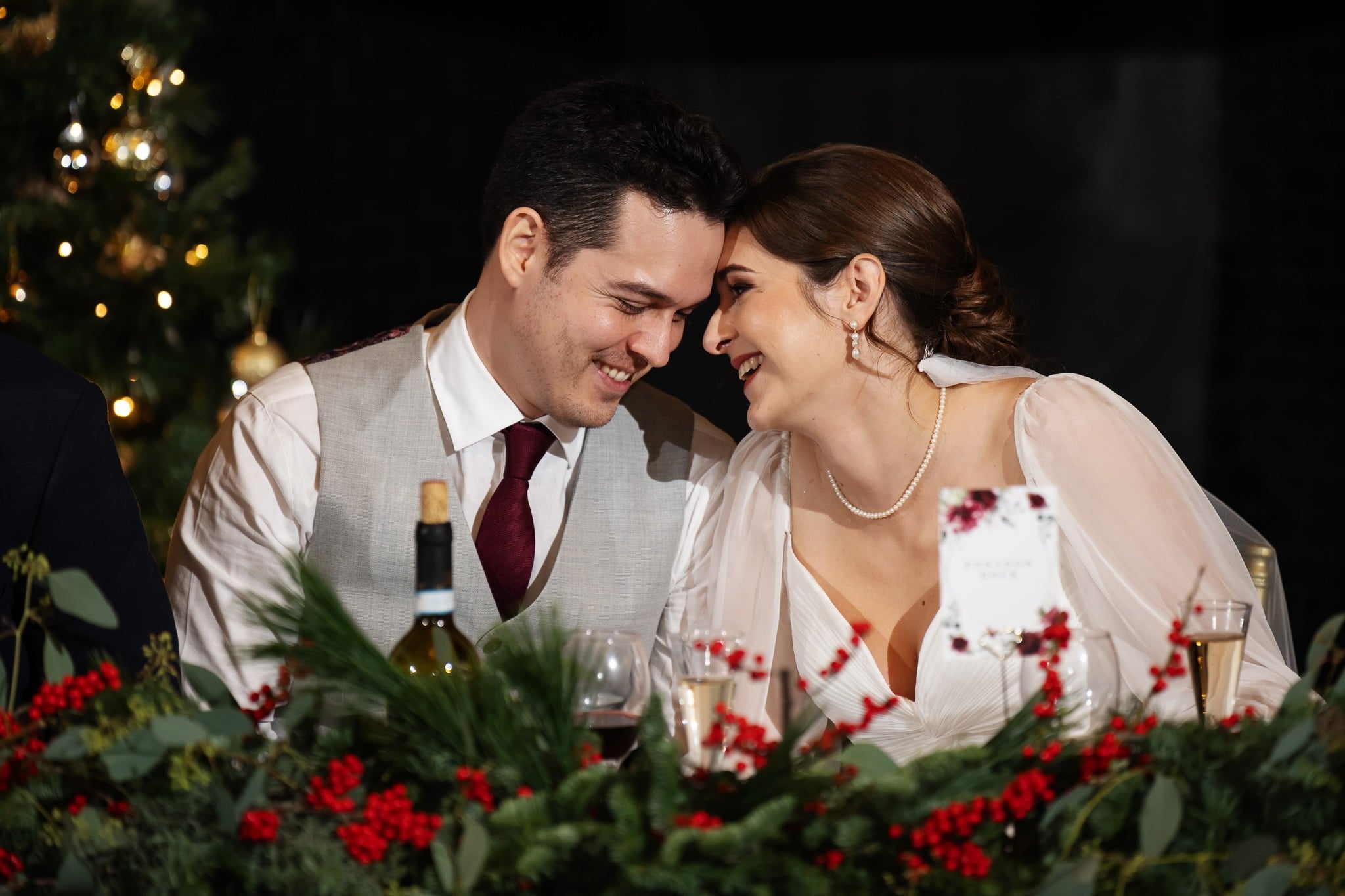 A couple in formal attire leans close with heads touching, smiling at a table adorned with festive greenery and red berries. A Christmas tree is in the background, capturing the warmth and elegance of their Surrey celebration, perfect for an Addington Palace wedding photography album.