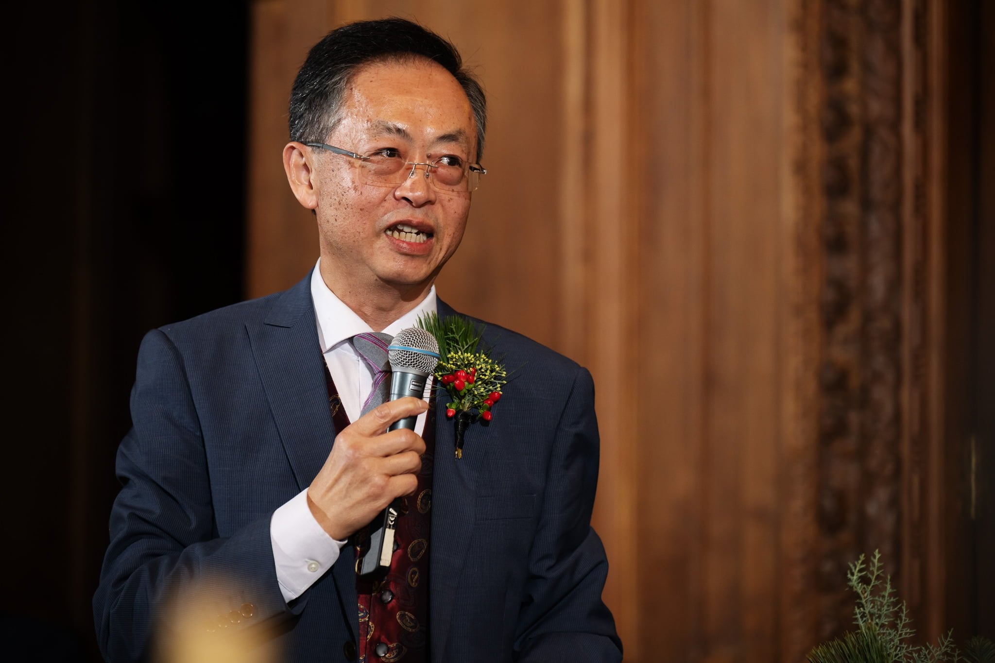 A man in a blue suit and glasses holds a microphone and speaks, a small bouquet pinned to his lapel. The setting is indoors with wooden decor, beautifully captured by an Addington Palace wedding photographer.