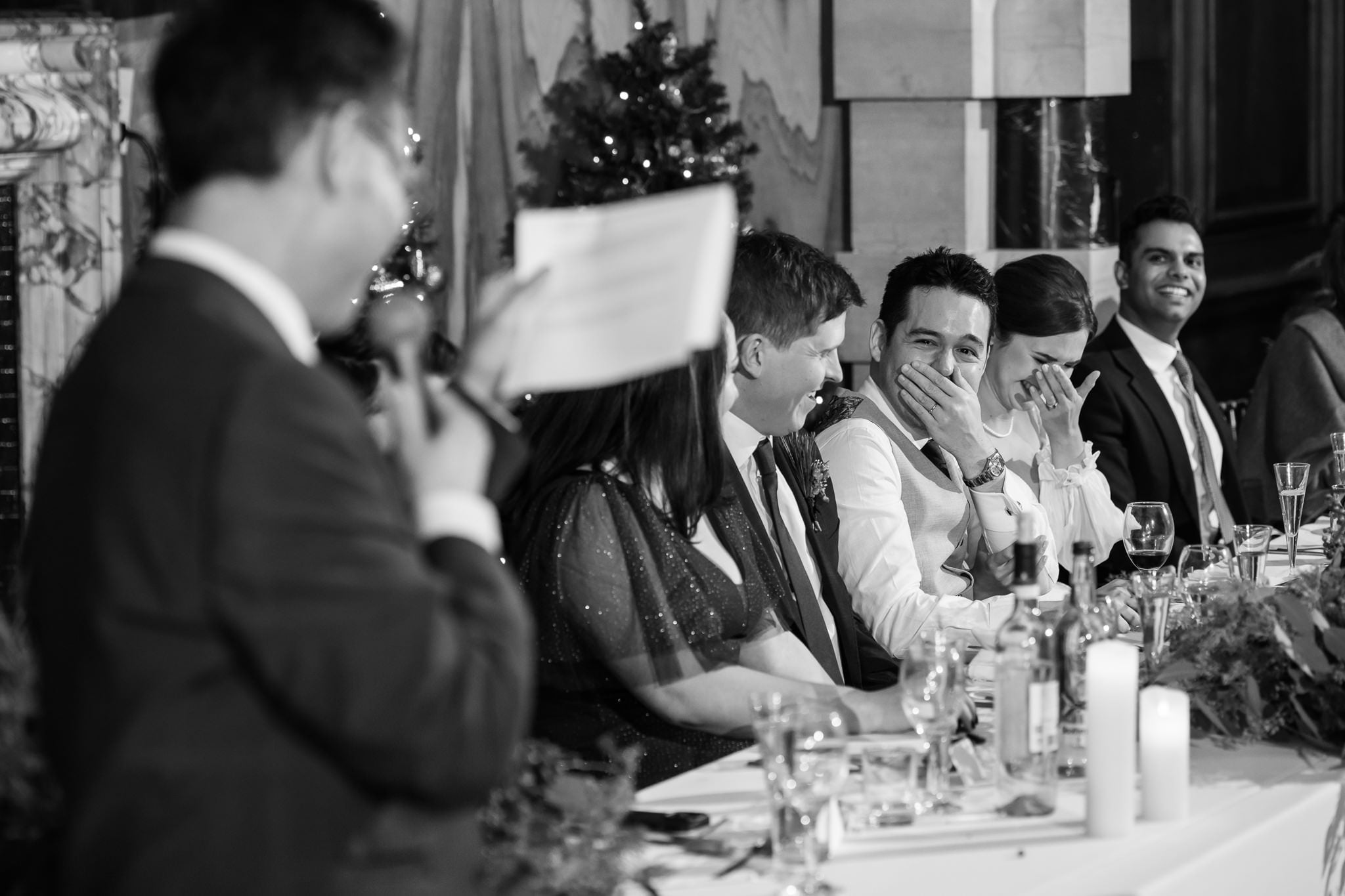 A man holds a microphone and piece of paper, speaking while five people seated at a decorated table laugh and smile during a formal event. This moment captures the joy and elegance often seen in Addington Palace wedding photography.