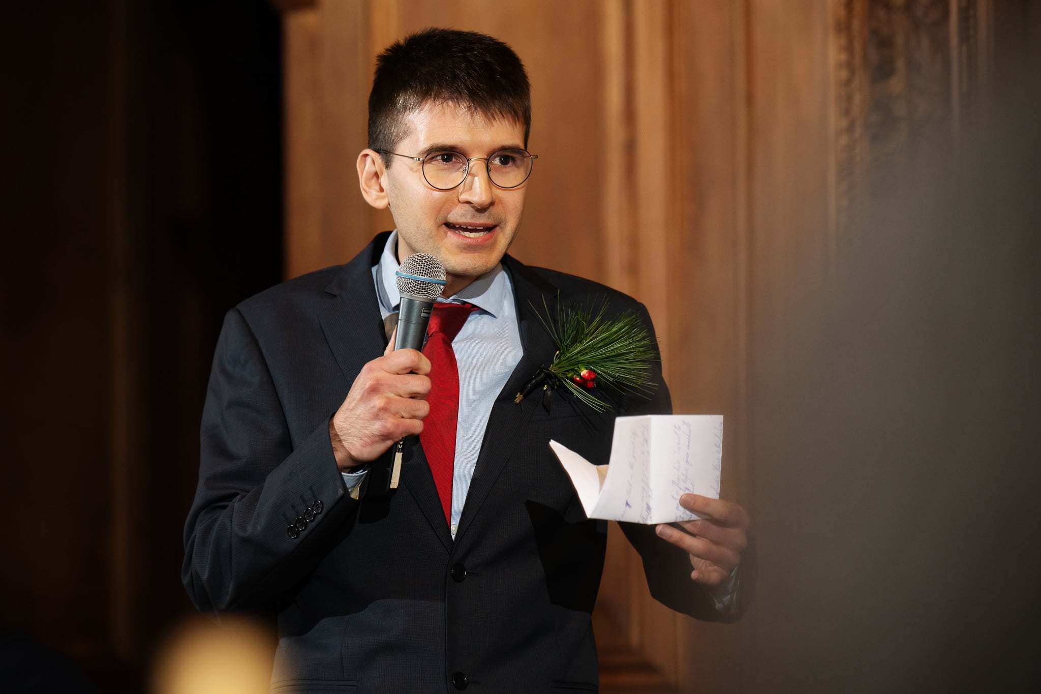 A man in a suit and glasses speaks into a microphone while holding a piece of paper. He has a decorative piece with greenery and a berry pinned to his suit. Wooden paneling is visible in the background, capturing the elegance typical of Addington Palace wedding photography in Surrey.