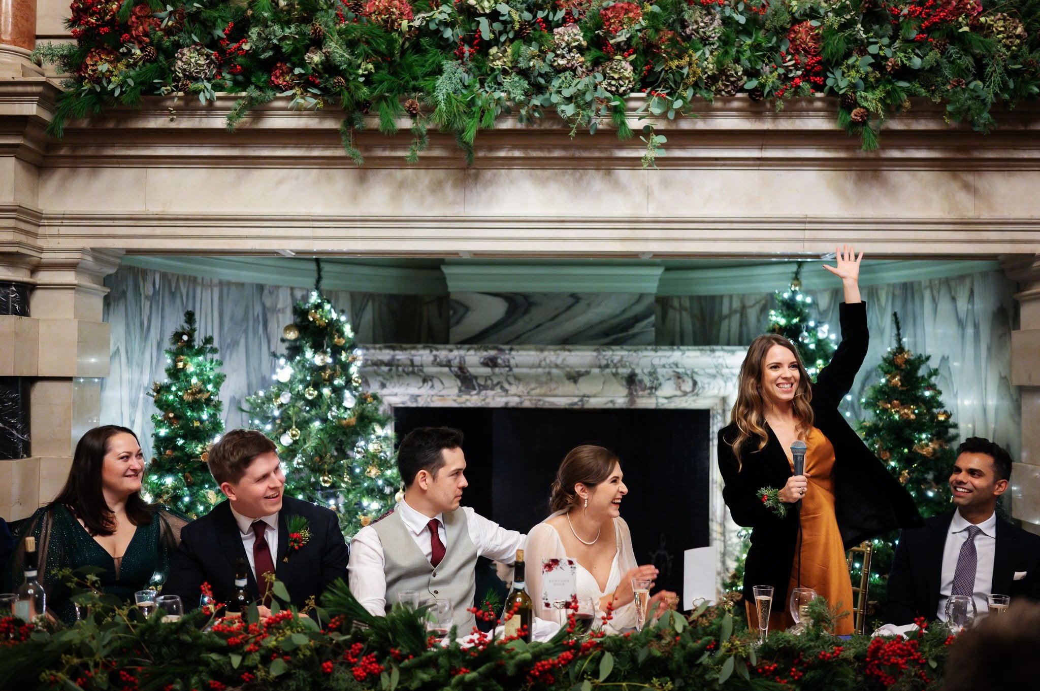 At a festive table adorned with greenery and flowers, five people are seated while a sixth stands, microphone in hand and raising one. Decorated Christmas trees and garlands form the backdrop, capturing the celebratory spirit of an unforgettable Addington Palace wedding in Surrey.