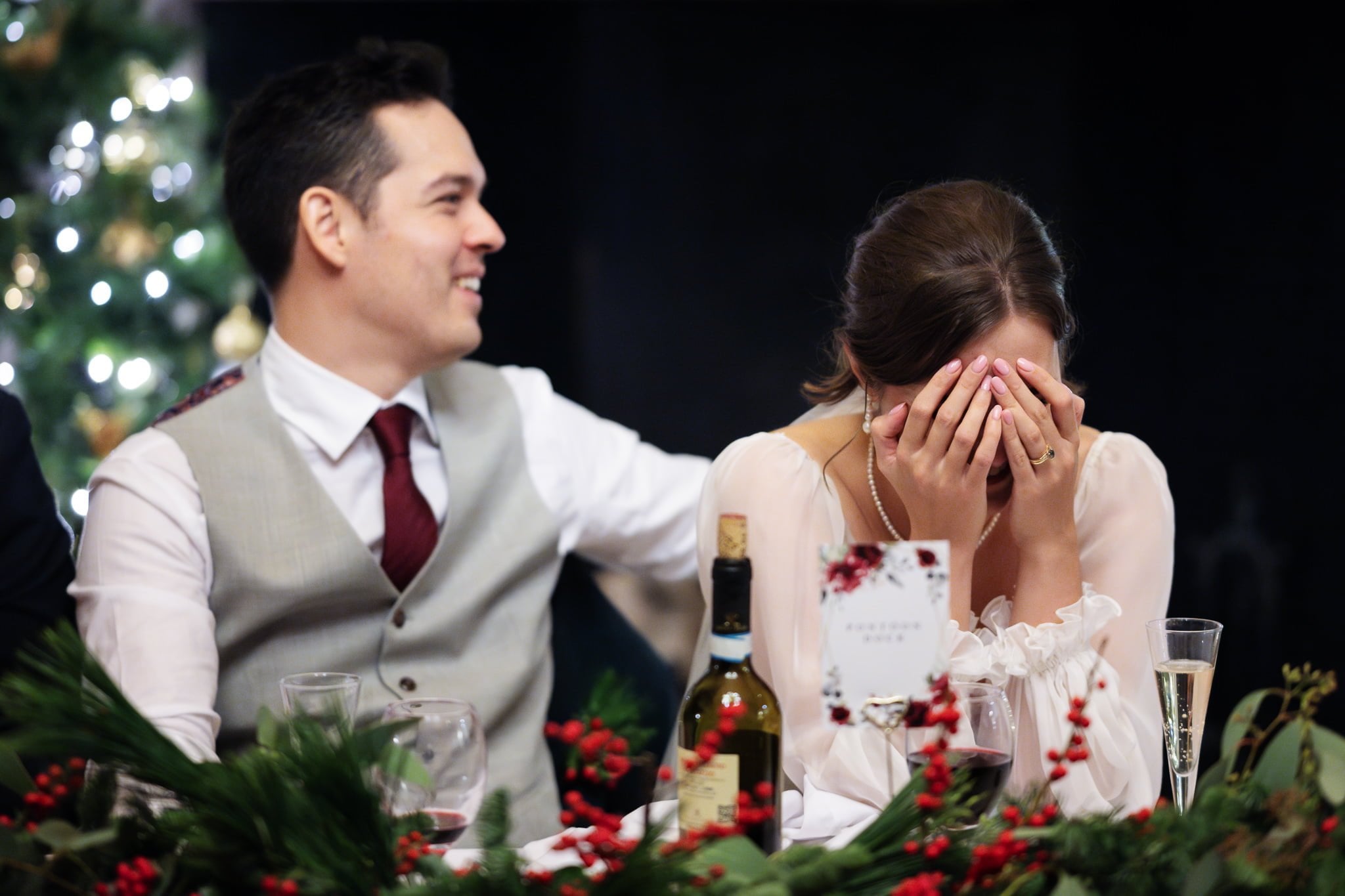 A woman covers her face with her hands while sitting beside a man in a vest and tie. They are sitting at a decorated table with festive greenery, wine, and glasses, perfectly captured by an Addington Palace wedding photographer.