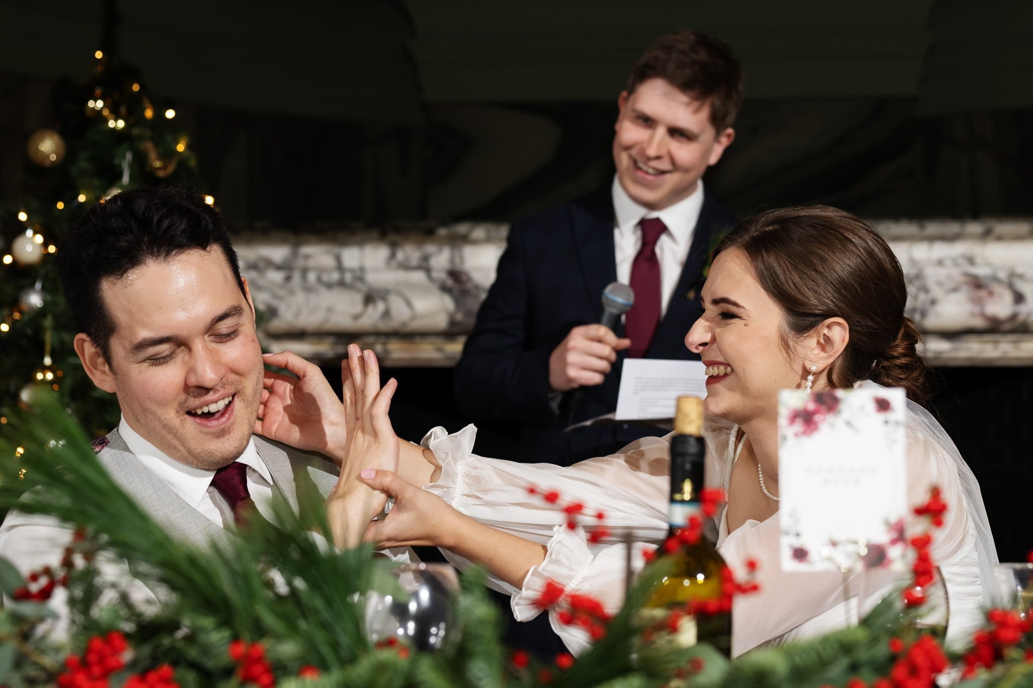 A woman playfully touches a man's cheek while he reacts with a smile. A man in a suit, holding a microphone, stands behind them. They are at a decorated table with festive greenery and red berries, all beautifully captured by the Addington Palace wedding photographer.