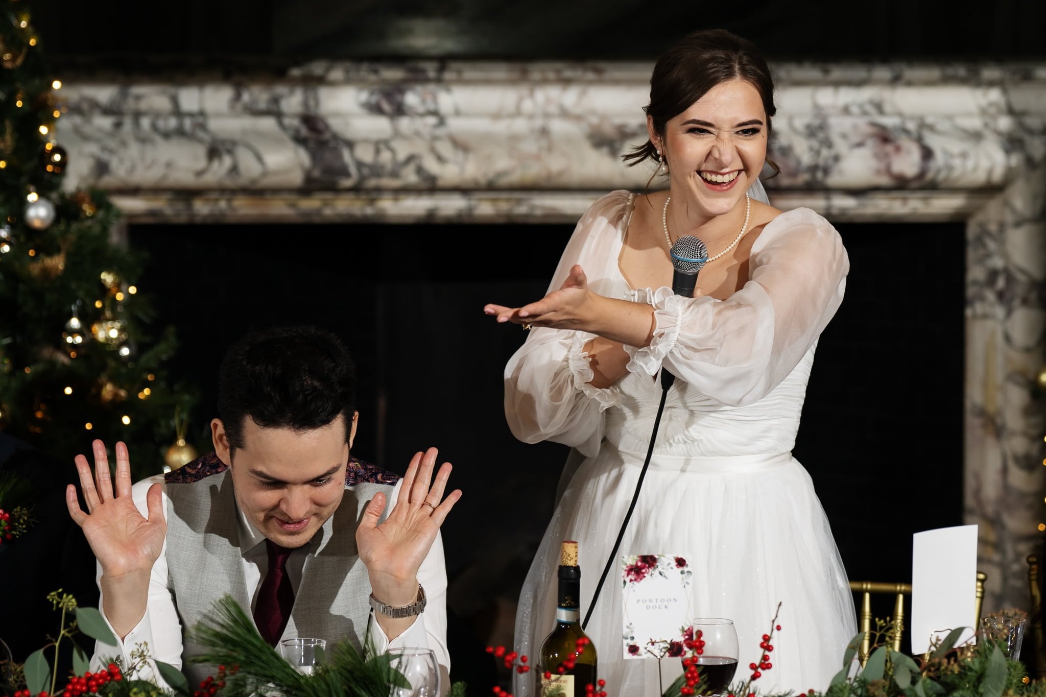 A bride holding a microphone and smiling gestures to the seated groom, who has his hands raised and eyes closed. They are at a beautifully decorated table at an enchanting Addington Palace wedding in Surrey.