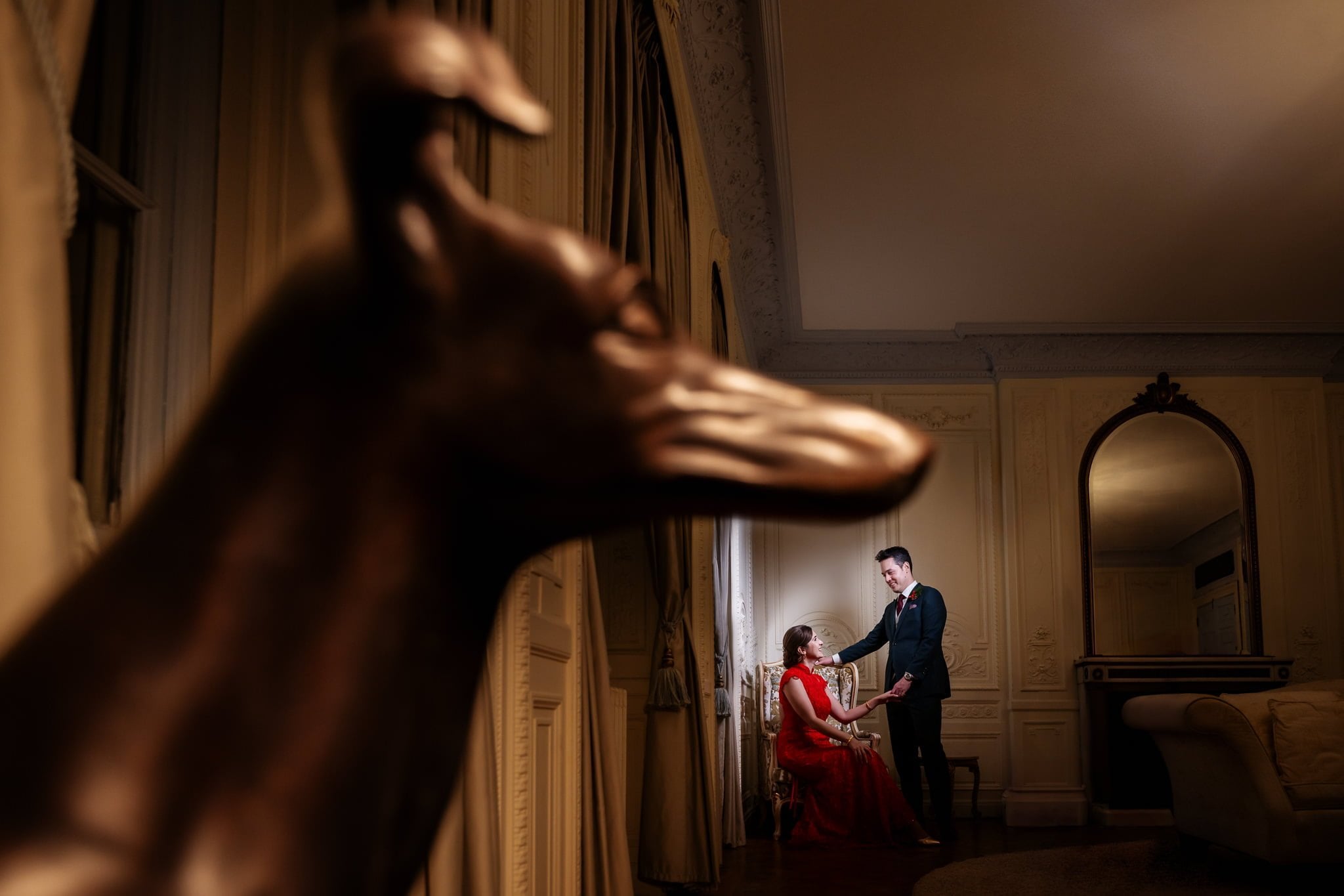 A couple dressed formally, with the woman seated and the man standing, share an intimate moment in an elegant room with a large mirrored wall and a prominent dog statue in the foreground—a perfect scene for Addington Palace wedding photography.