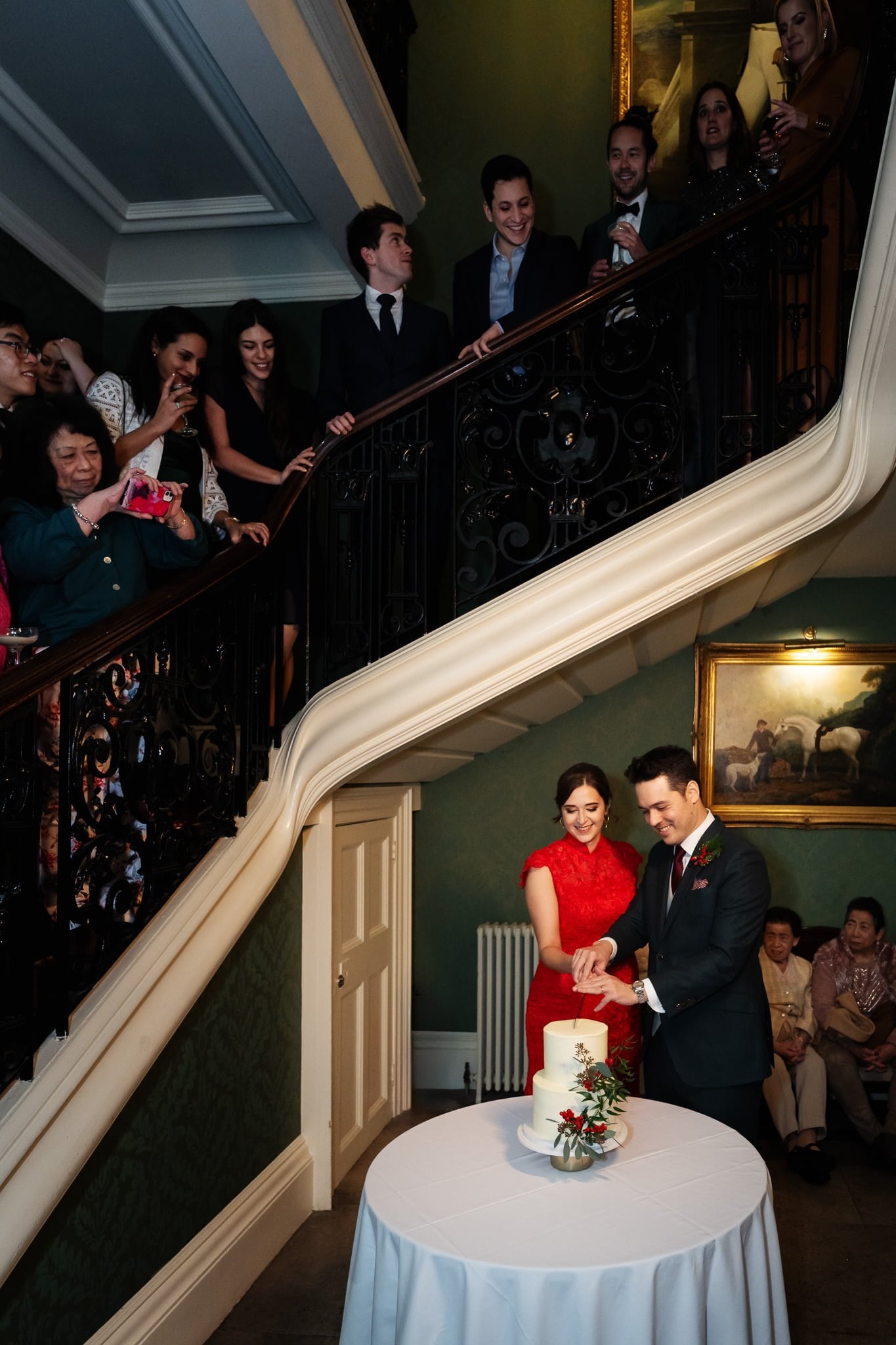 A couple in formal attire cuts a wedding cake placed on a round table, perfectly captured by an Addington Palace wedding photographer. Guests stand on a staircase behind them, watching and taking pictures.