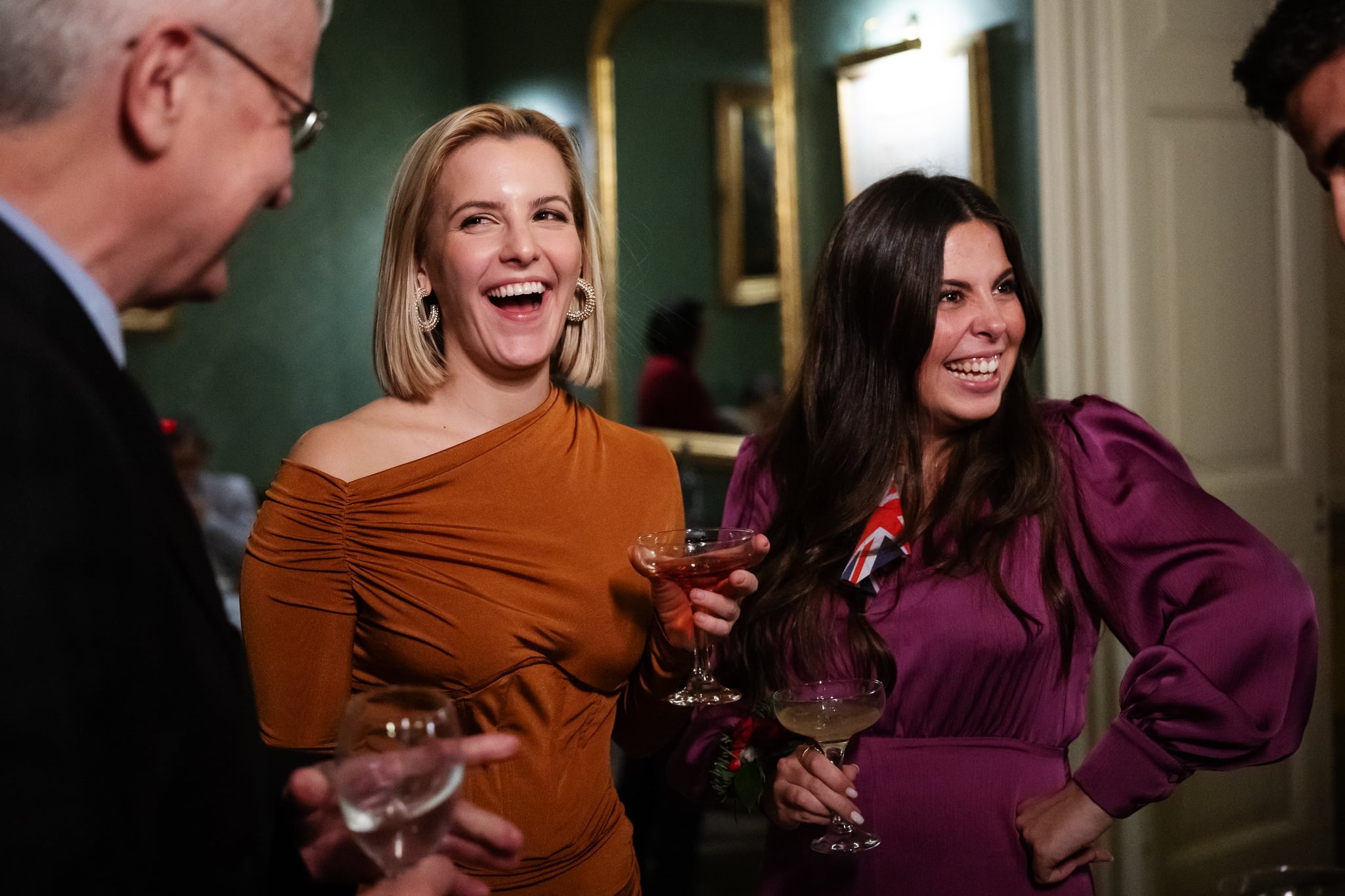 Two women in elegant dresses laugh while holding cocktails at a social gathering at Addington Palace. A man in a suit is partially visible on the left, and a fourth person is on the right, all captured beautifully by an Addington Palace wedding photographer.