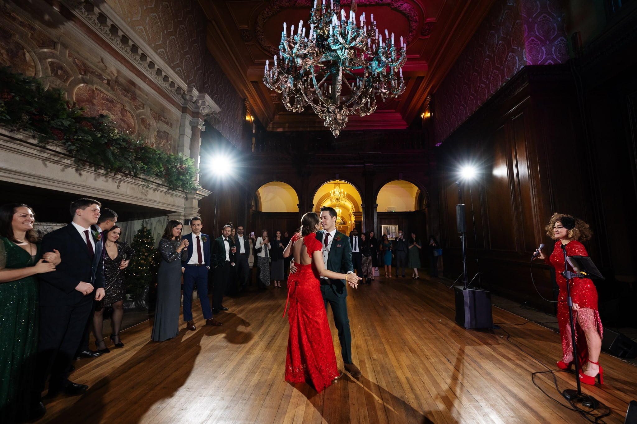 A couple dances in the center of an elegant ballroom at their Addington Palace wedding, with guests watching. The room features a large chandelier, ornate wall decorations, and wooden flooring. A musician in red performs on the right. Perfect for capturing timeless Addington Palace wedding photography.
