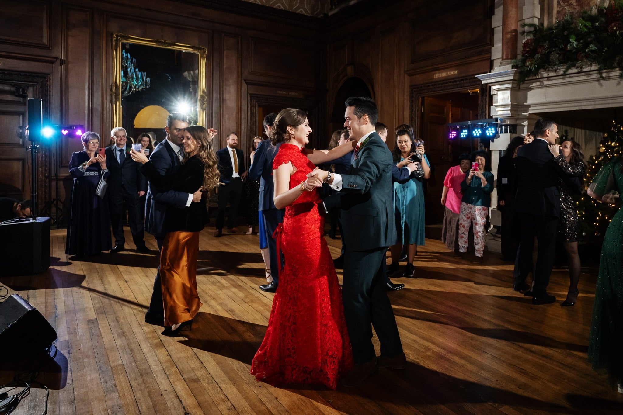 People dancing in an elegant, wood-paneled room with festive decorations at a stunning Addington Palace wedding in Surrey. The women in red and green dresses are prominently featured while couples dance gracefully in the background.