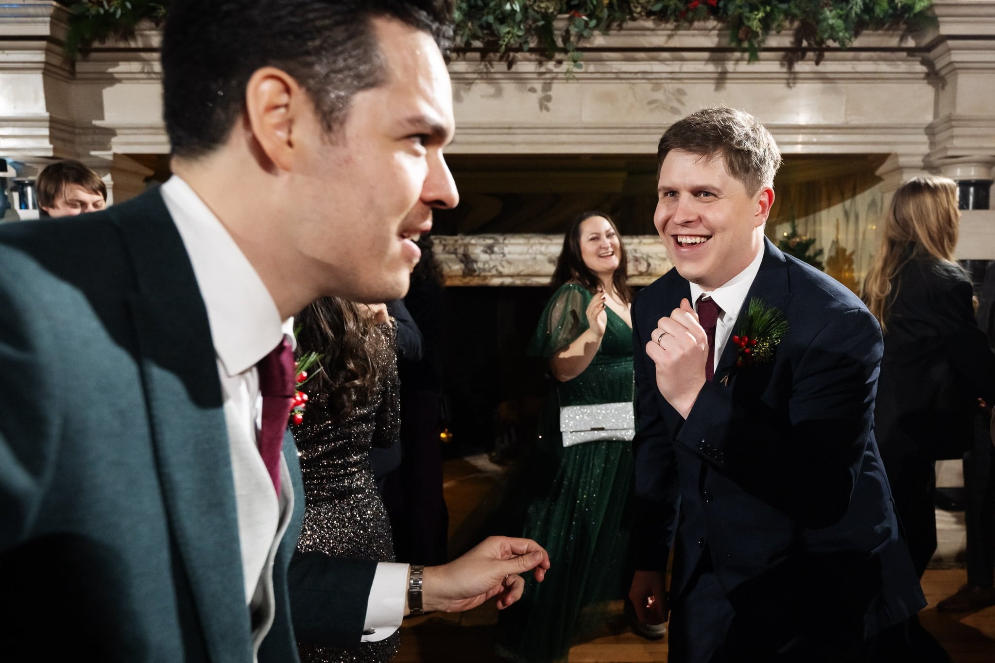 Two men in suits engage in conversation at a social gathering, with a woman in a green dress and others in the background—a perfect moment captured by an Addington Palace wedding photographer during a Surrey celebration.