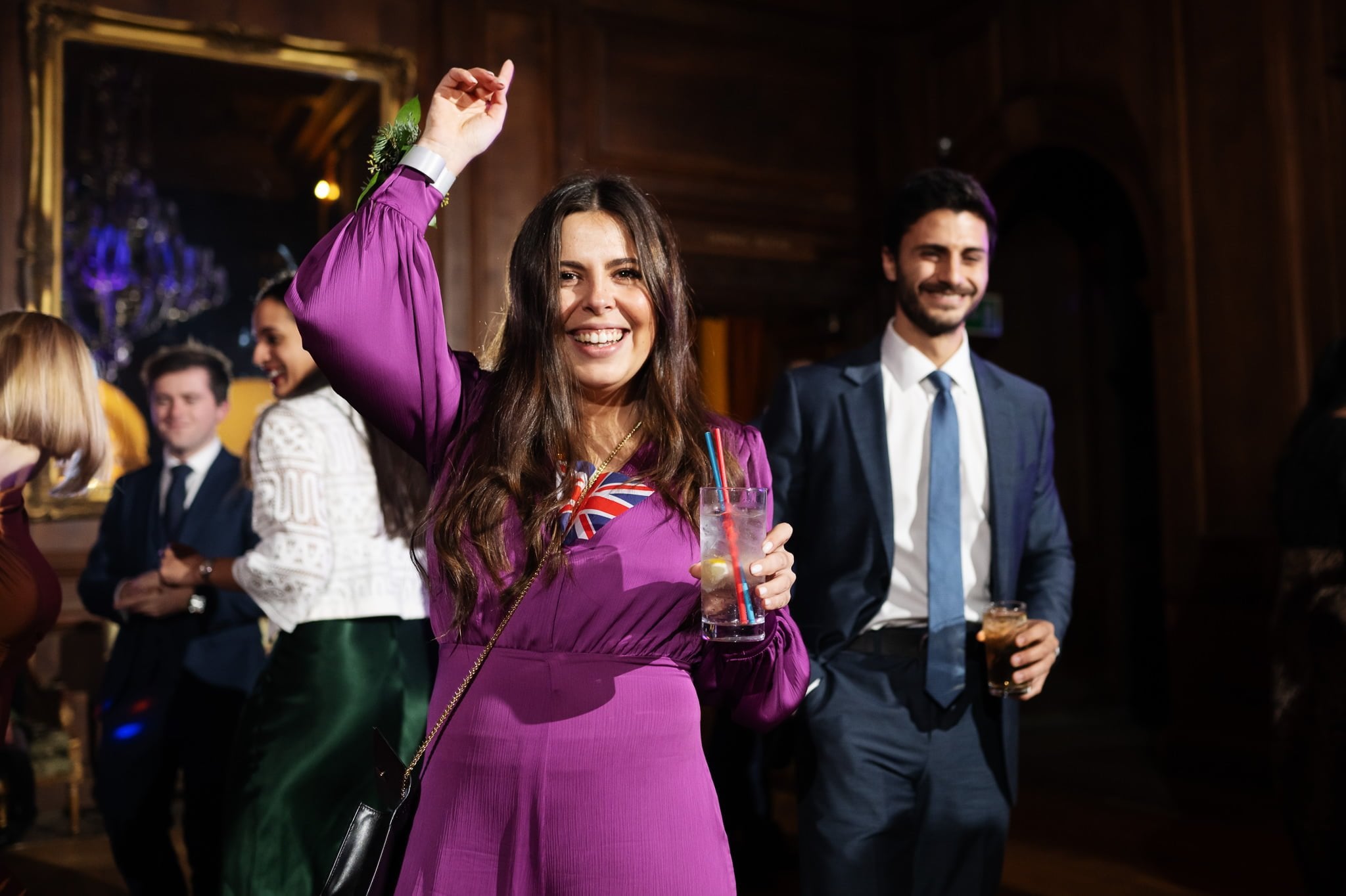 A woman in a purple dress smiles and raises her arm while holding a drink, surrounded by other people in formal attire at a festive Addington Palace wedding in a dimly lit wooden room.