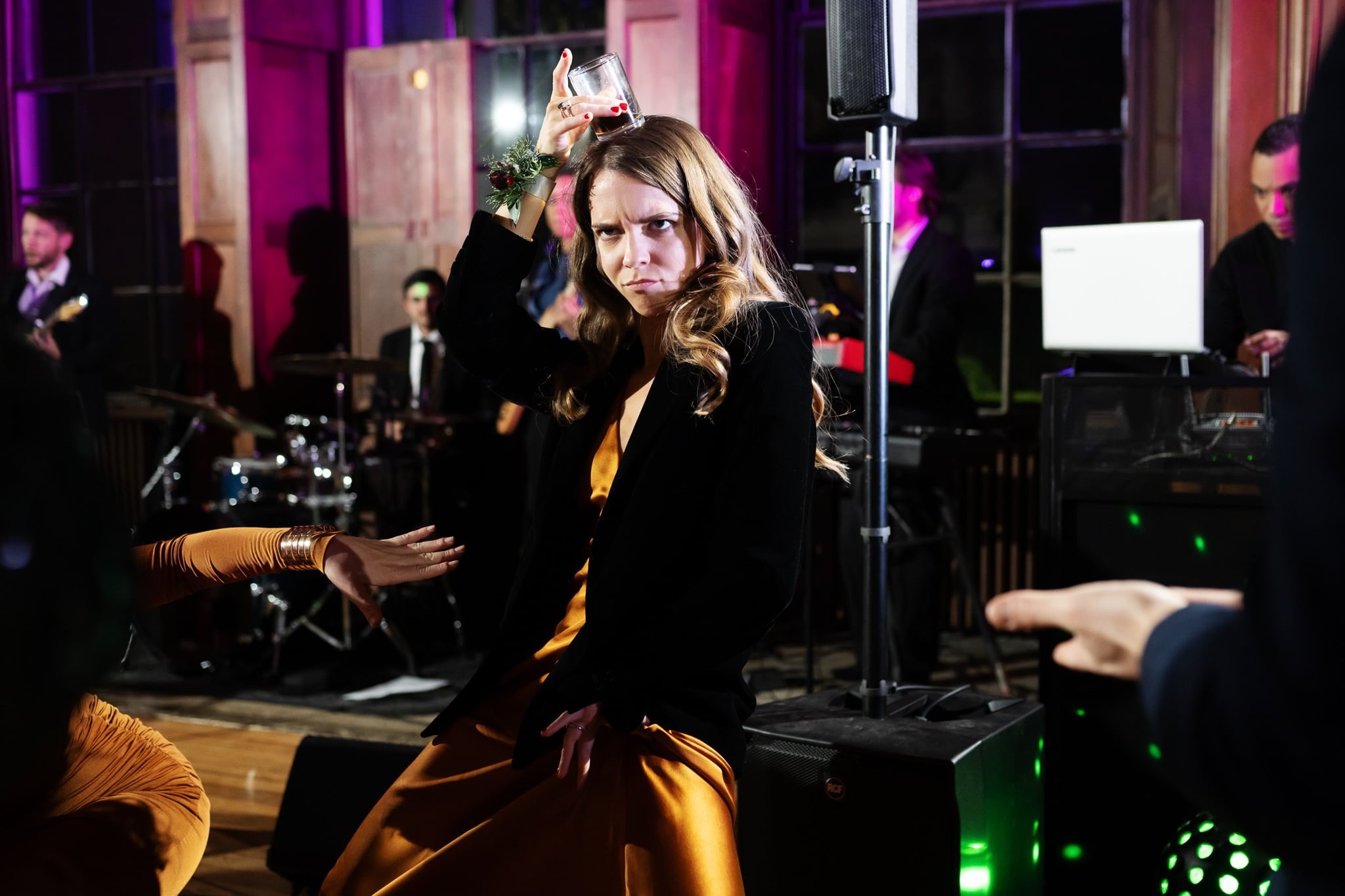 A woman in a fancy outfit holds a drink above her head while dancing at a lively indoor wedding with a band playing in the background, captured perfectly by an Addington Palace wedding photographer.