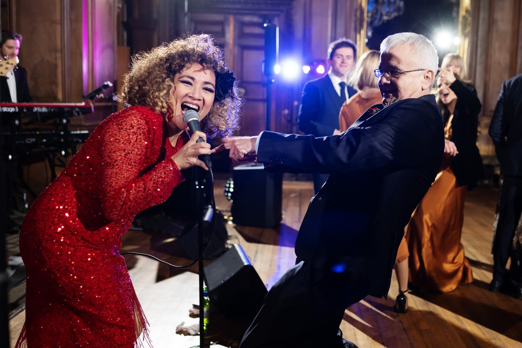 A woman in a red dress sings into a microphone while a man in a suit dances energetically beside her, capturing the joyous spirit of an Addington Palace wedding in Surrey.