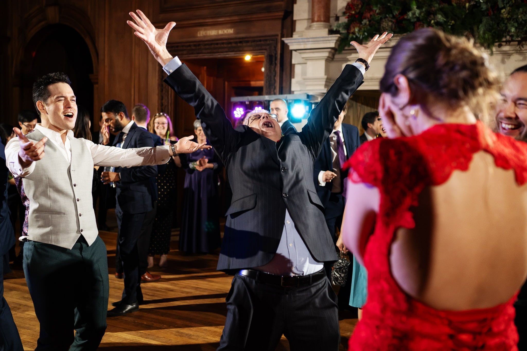 People are dancing and cheering joyfully at an indoor event. A man in a suit stands in the center with arms raised, while others around him are smiling and clapping, capturing the exuberance of an unforgettable Addington Palace wedding.