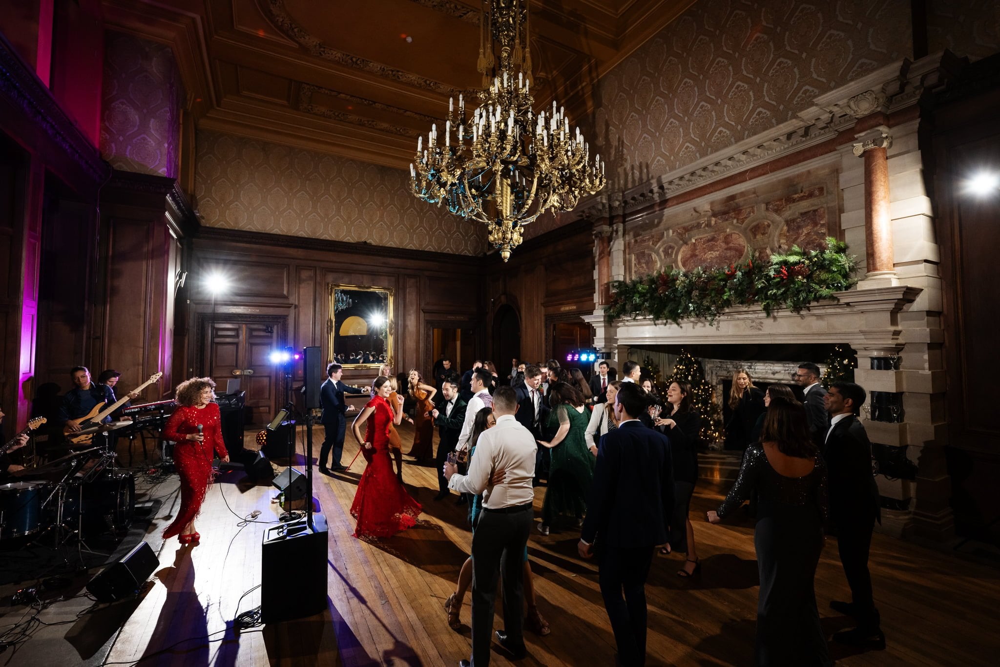 A large group of people in formal attire dance under a grand chandelier in an ornately decorated ballroom at a Surrey wedding, with a live band performing on stage.