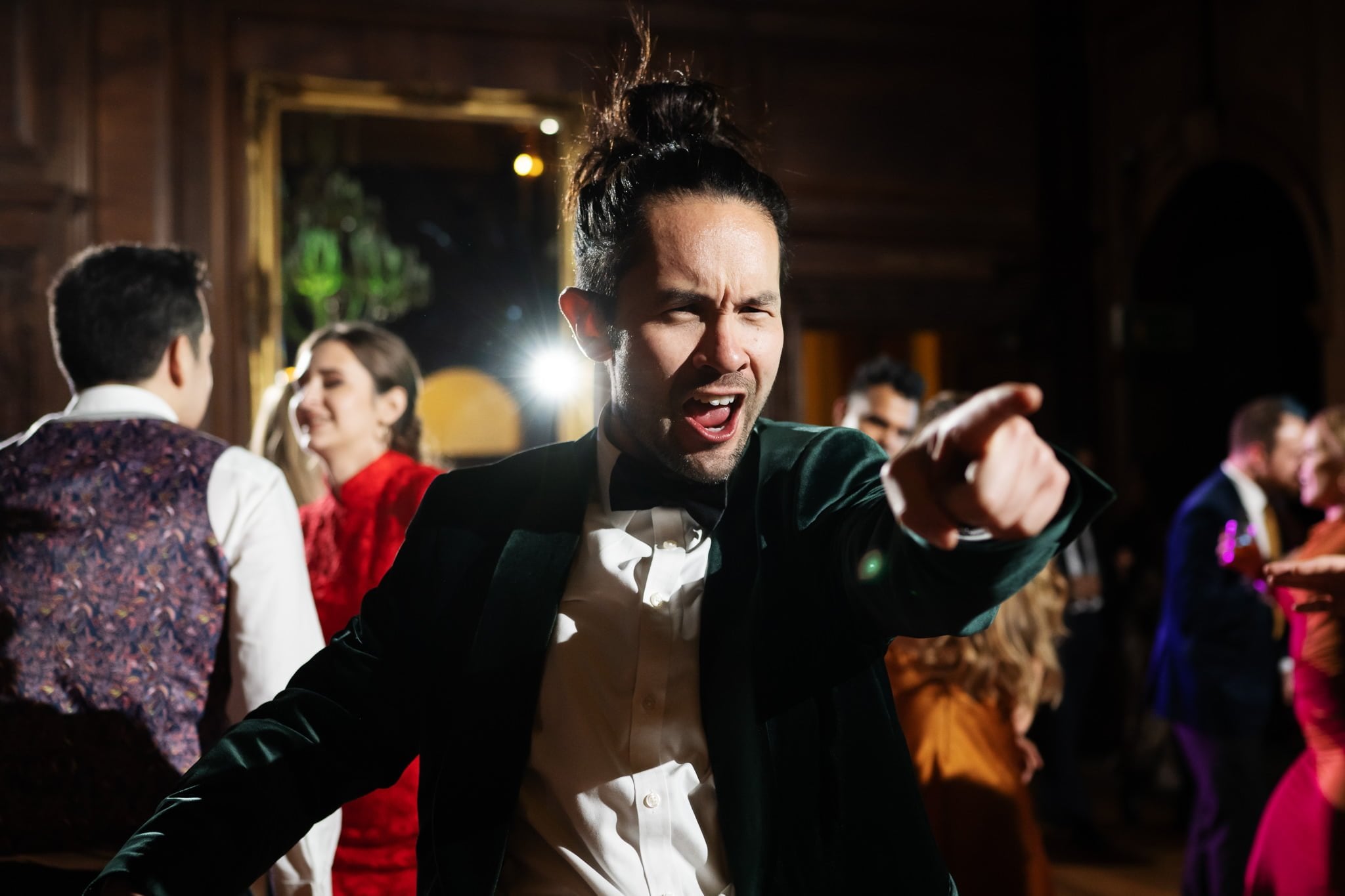 A man in formal attire with a bowtie enthusiastically points at the camera while dancing at an indoor event at Addington Palace, with other people in the background. Perfect for immortalizing vibrant moments, this could be part of your Surrey wedding photography collection.