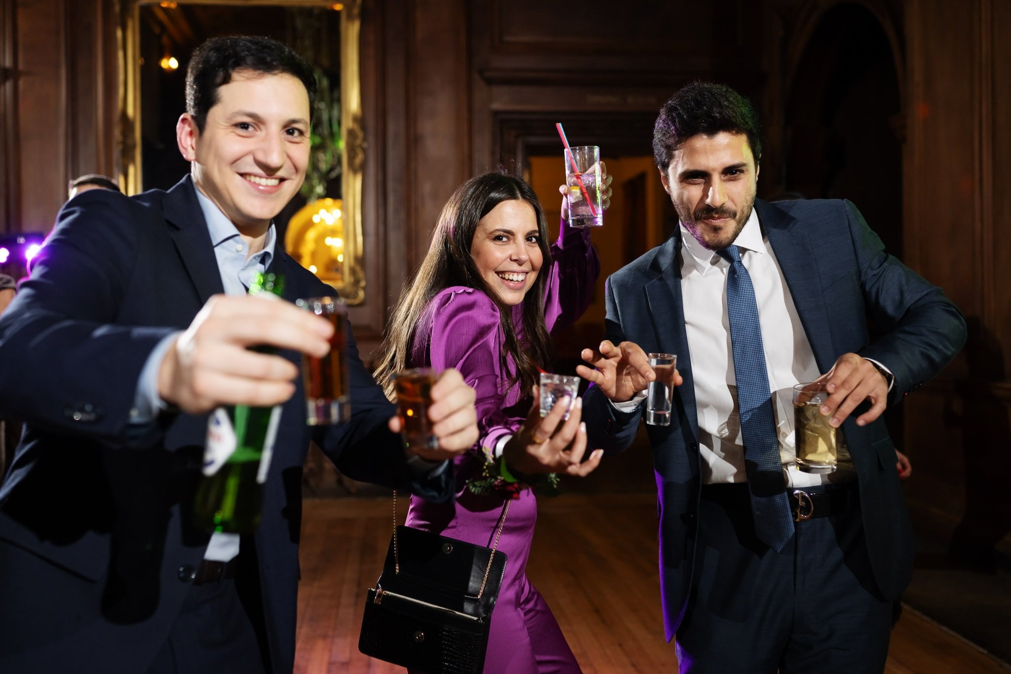 Three people dressed in formal attire are smiling and holding up drinks in a dimly lit, elegant room with wooden walls and a mirror, capturing the joyous essence of an Addington Palace wedding.