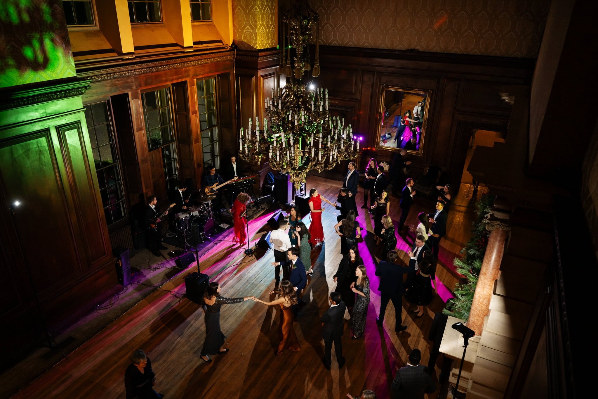 An overhead view of people dancing in a large, elegant hall at Addington Palace wedding with a live band performing; chandeliers and wooden paneling adorn the space.