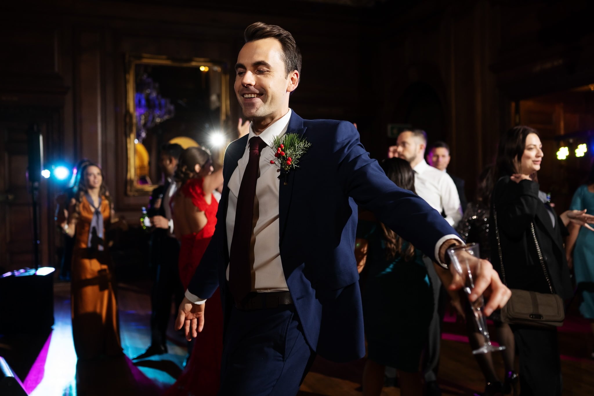 A man in a blue suit, holding a drink, smiles and walks confidently at an Addington Palace wedding with people in the background.