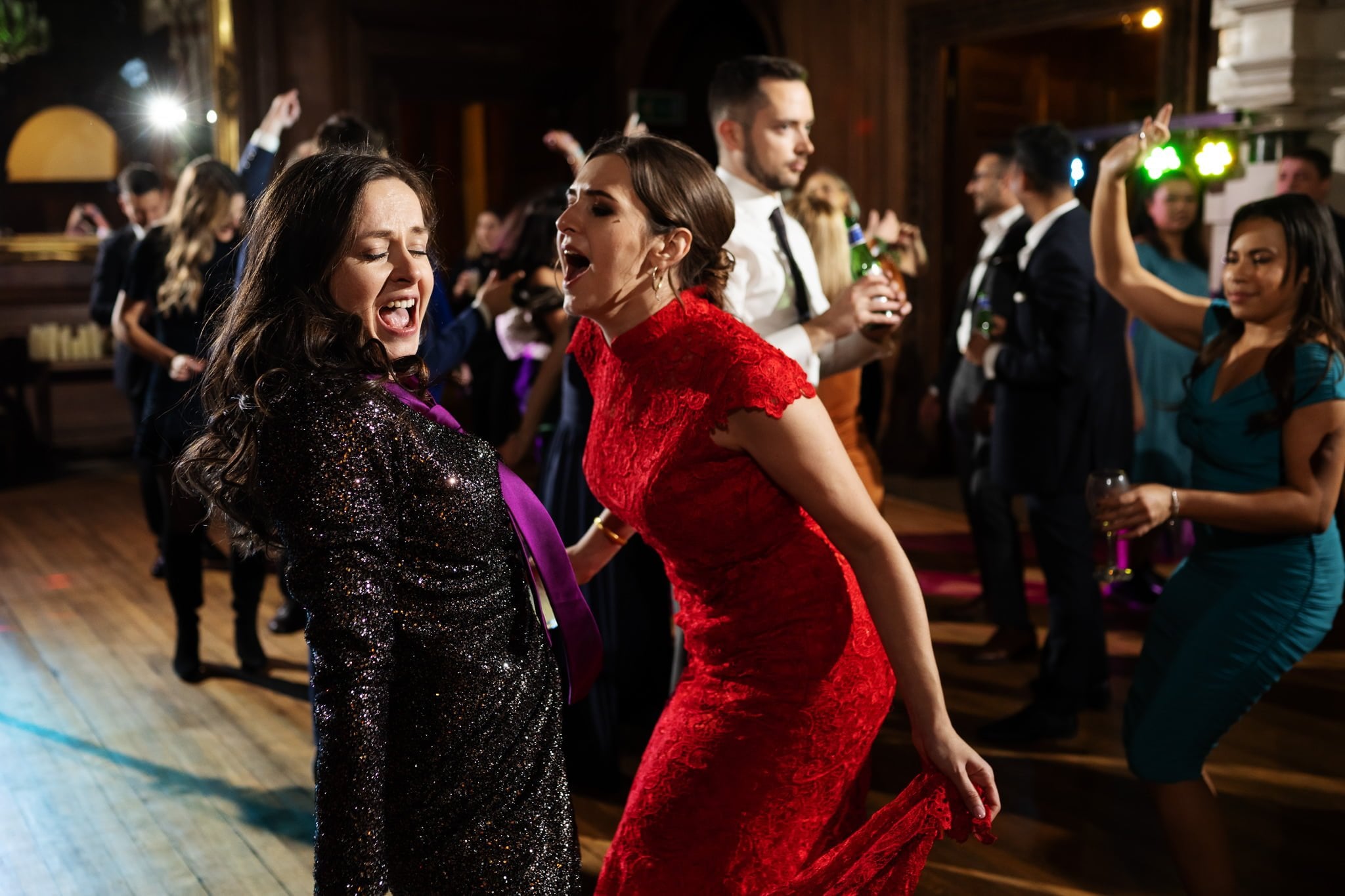 Two women in formal dresses dance energetically at a lively Addington Palace wedding with a crowd of people in the background. One woman wears a black sequined dress, and the other wears a red dress. Capture this vibrant moment through the lens of an Addington Palace wedding photographer in Surrey.