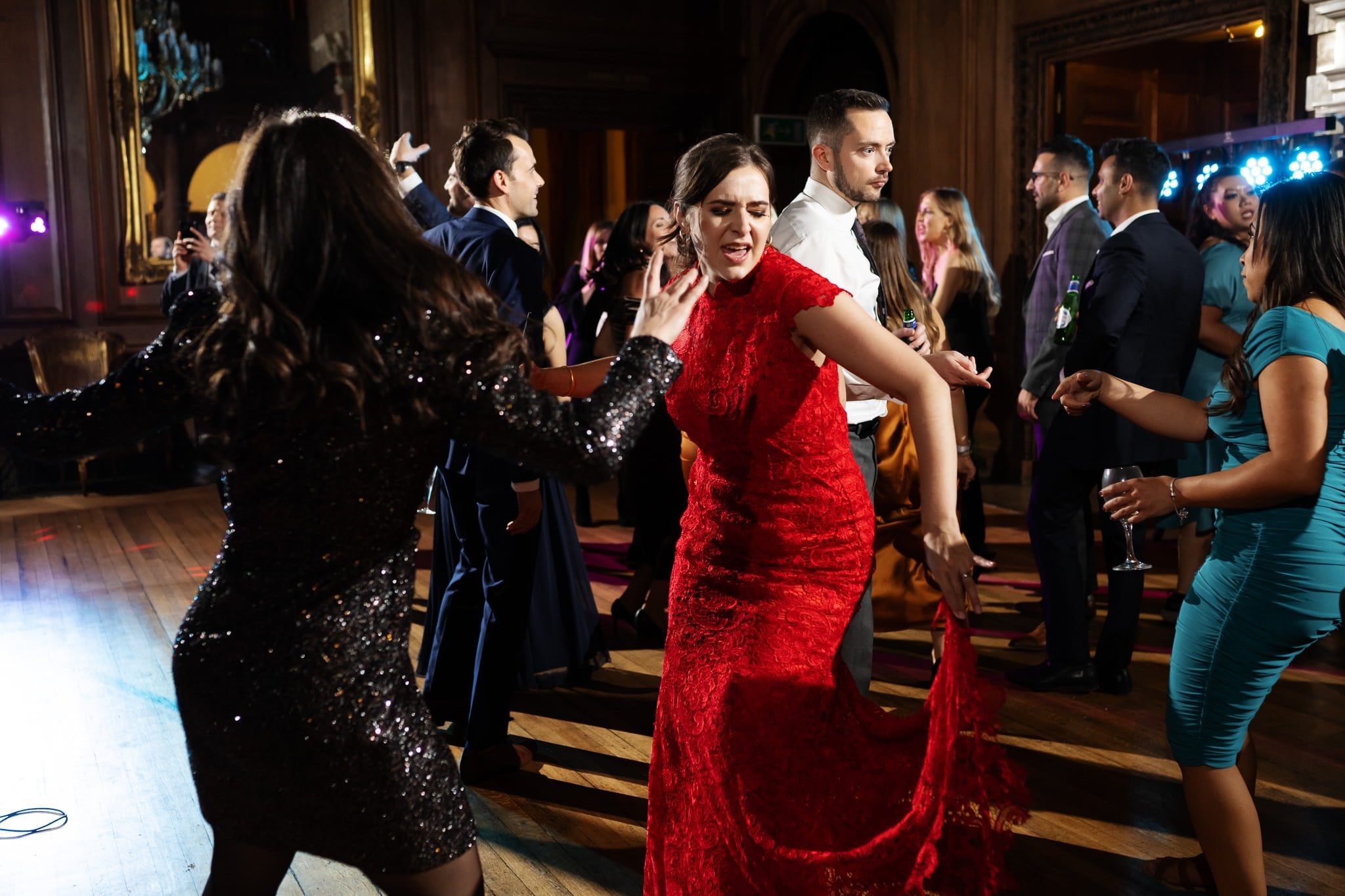 At an elegant indoor event, people are dancing with joy; a woman in a red dress and another in a black sequin dress grace the foreground. This scene could easily be captured by an Addington Palace wedding photographer at a stunning Surrey wedding.