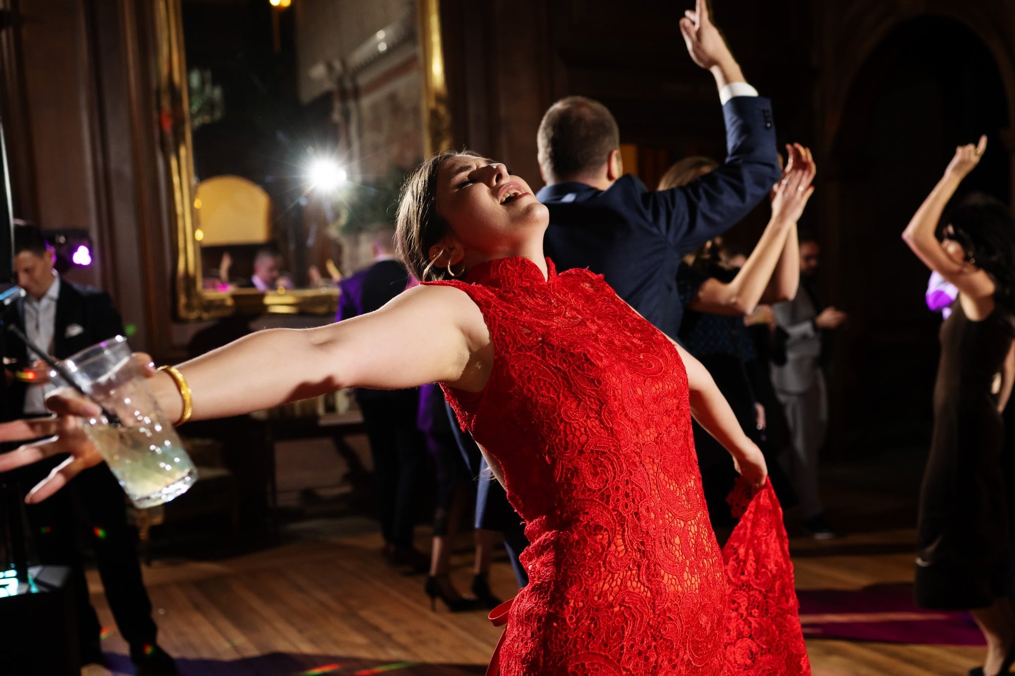 A woman in a red dress dances with her arms outstretched, holding a drink in one hand. Several people are dancing in the background of this dimly lit room with wooden floors and a large mirror, captured perfectly by an Addington Palace wedding photographer at the Surrey reception.