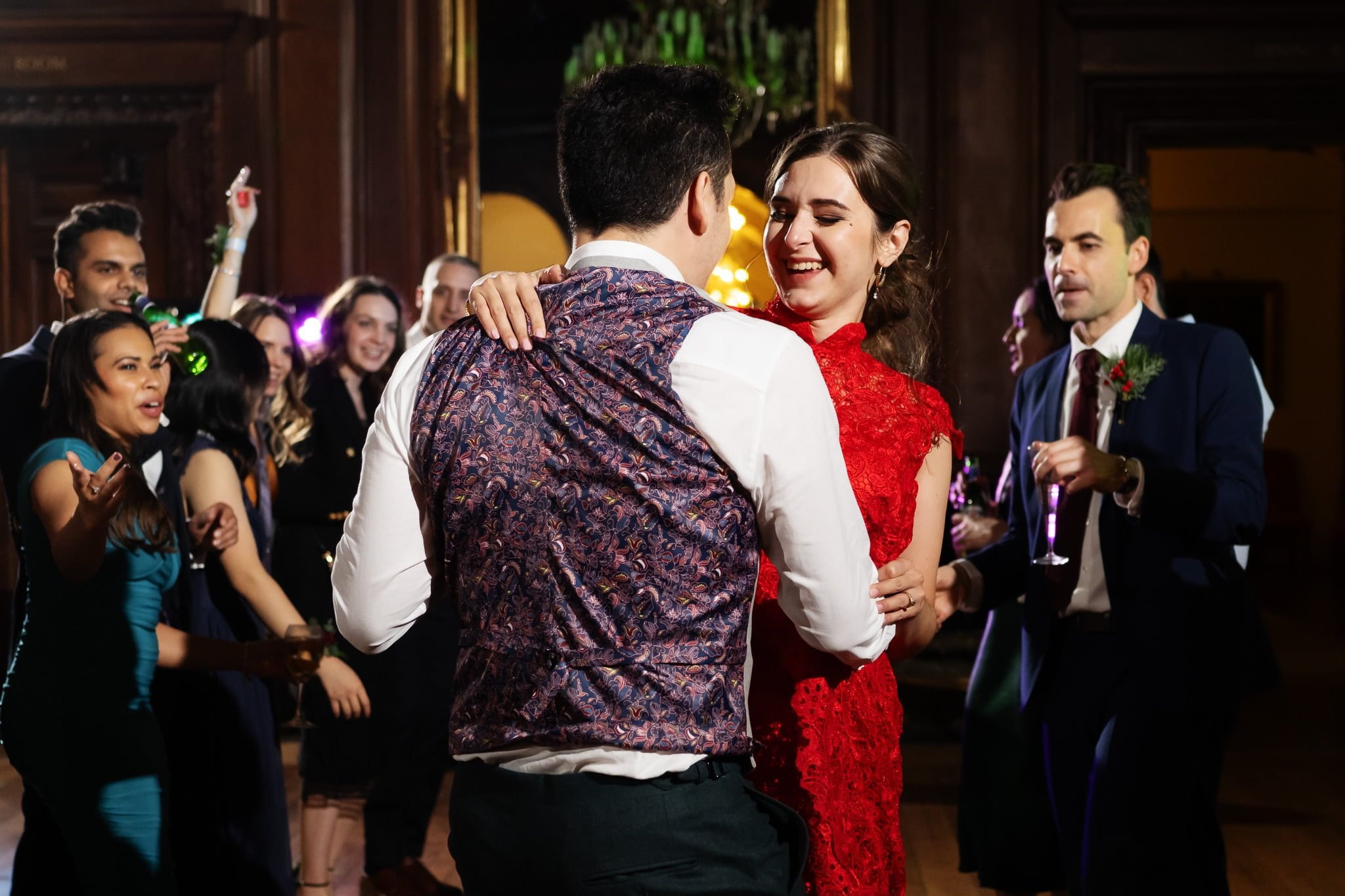 A man and woman dance together in the center of a room, surrounded by other people who appear to be celebrating at an Addington Palace wedding. The woman wears a red dress, and the man, with his back to the camera, is wearing a patterned vest.