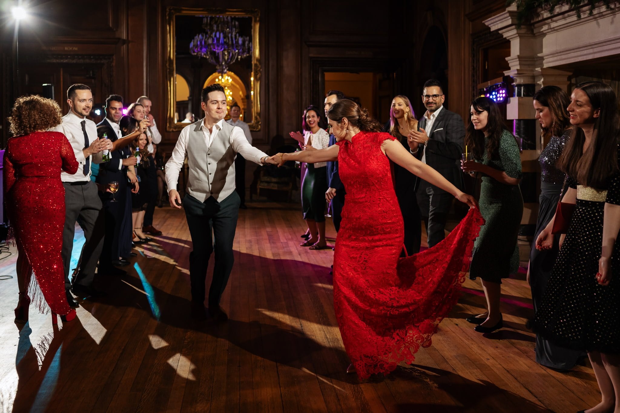A man and woman dressed formally dance in the center of a wooden floor with others clapping and watching in an elegant, dimly lit room, perfectly capturing the ambiance of a Surrey wedding.