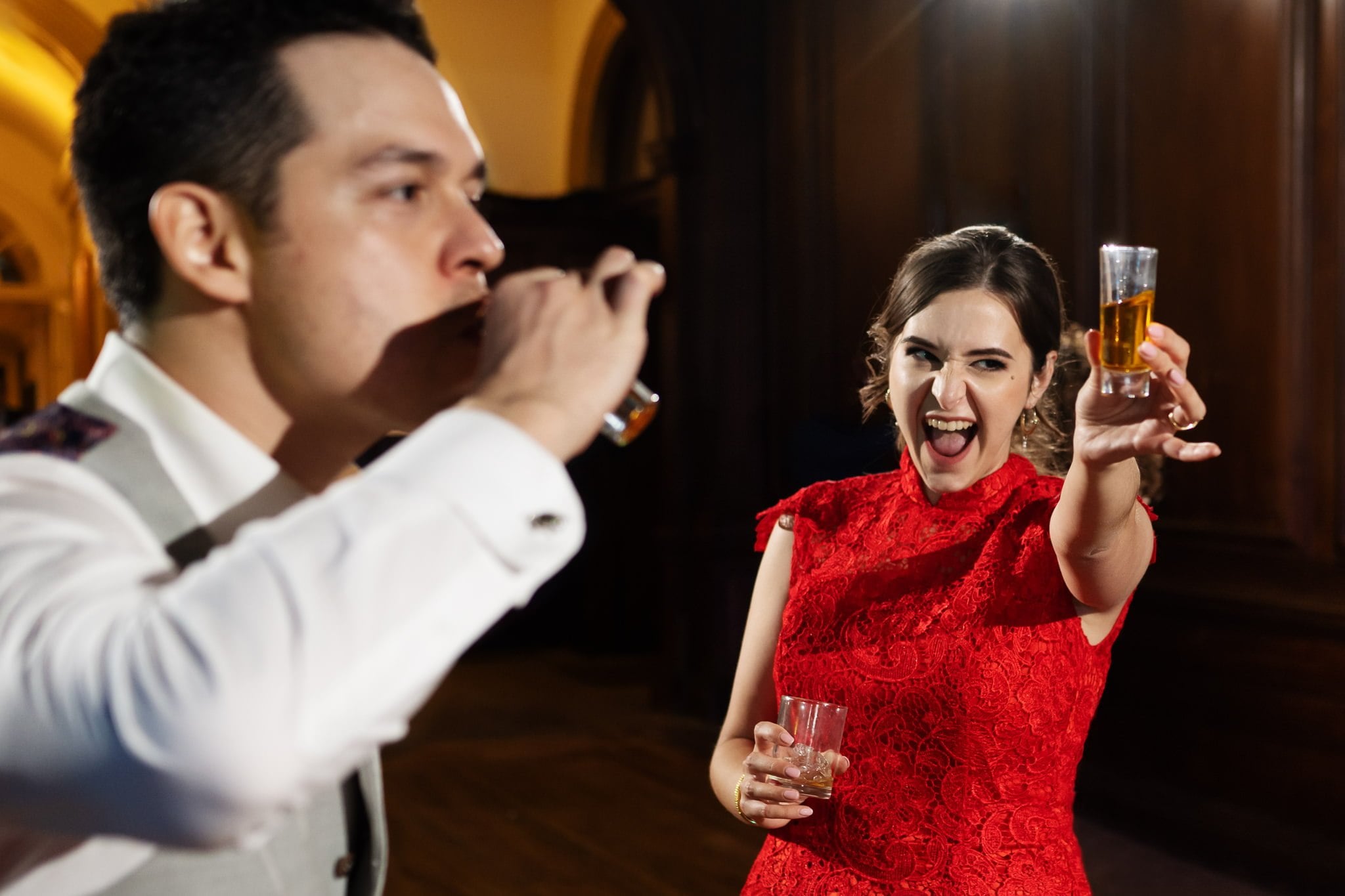 A woman in a red dress holds up a glass of amber liquid, smiling, while a man in a white shirt beside her drinks from a glass during an Addington Palace wedding.