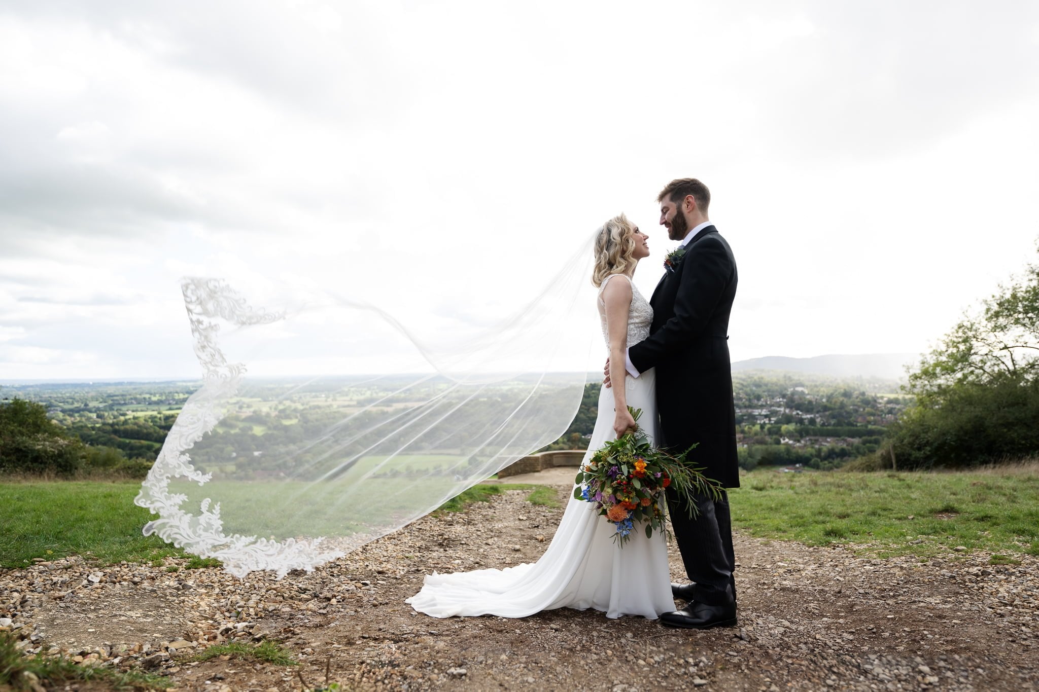 A bride and groom standing on a dirt path with their veil blowing in the wind.