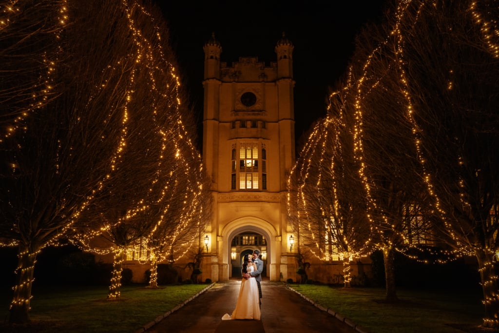 A couple stands under an illuminated archway at night, framed by rows of trees adorned with twinkling lights, in front of Danesfield House Wedding venue.