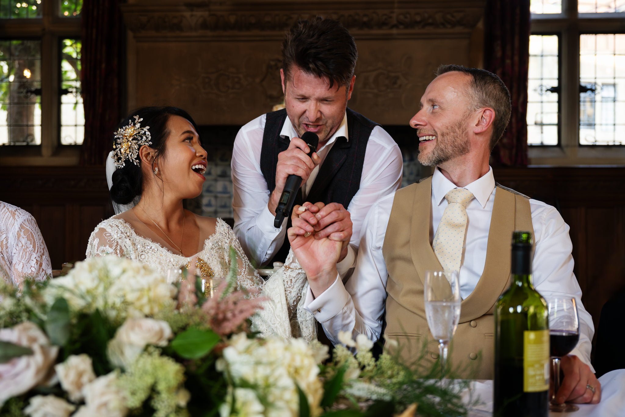 A wedding scene from a Fanhams Hall wedding. A bride and groom seated at a table, laughing and holding hands, while a surprise singing waiter stands between them singing.