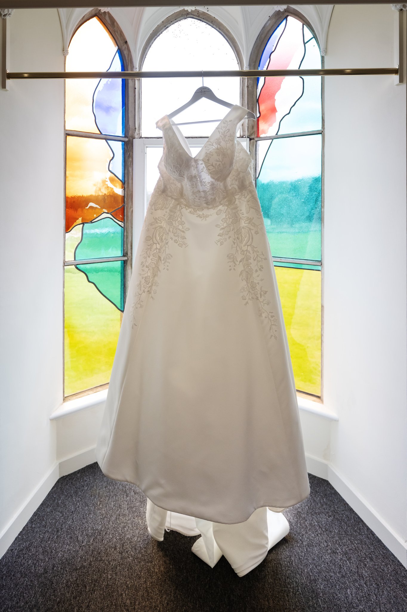 A white wedding dress hangs on a hanger in front of a stained-glass window with colorful designs, capturing the timeless elegance of Liz & Olly's wedding at Missenden Abbey.
